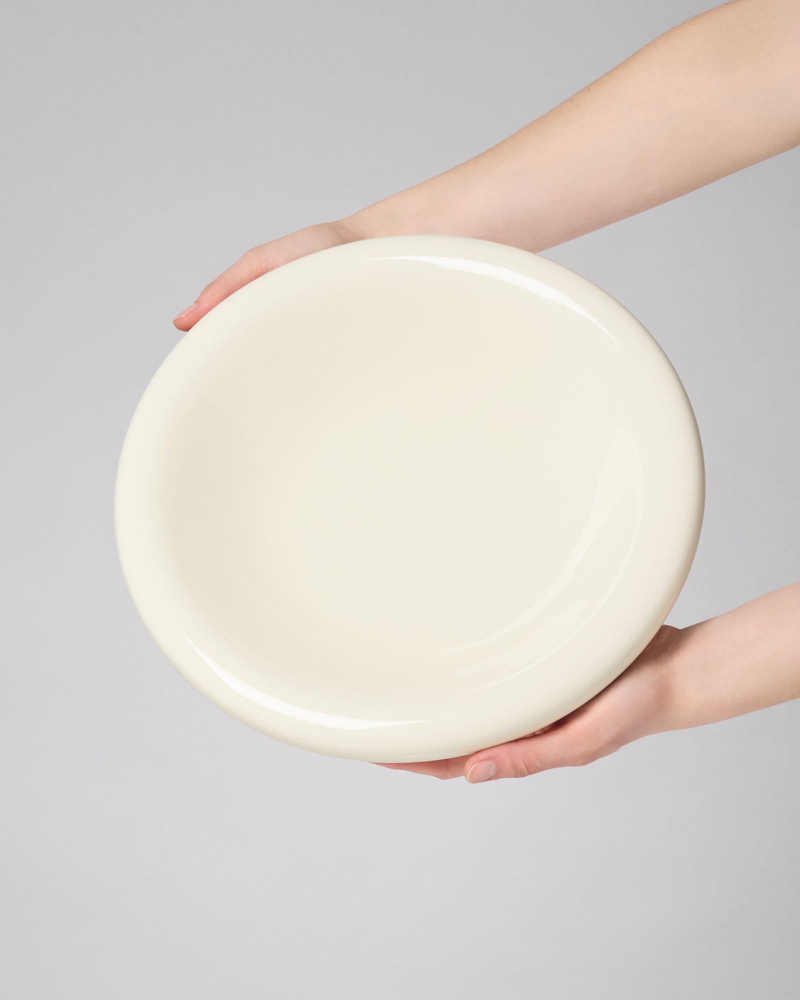 Zwei Hände halten eine große, cremefarbene, runde Schüssel vor einem grauen Hintergrund.