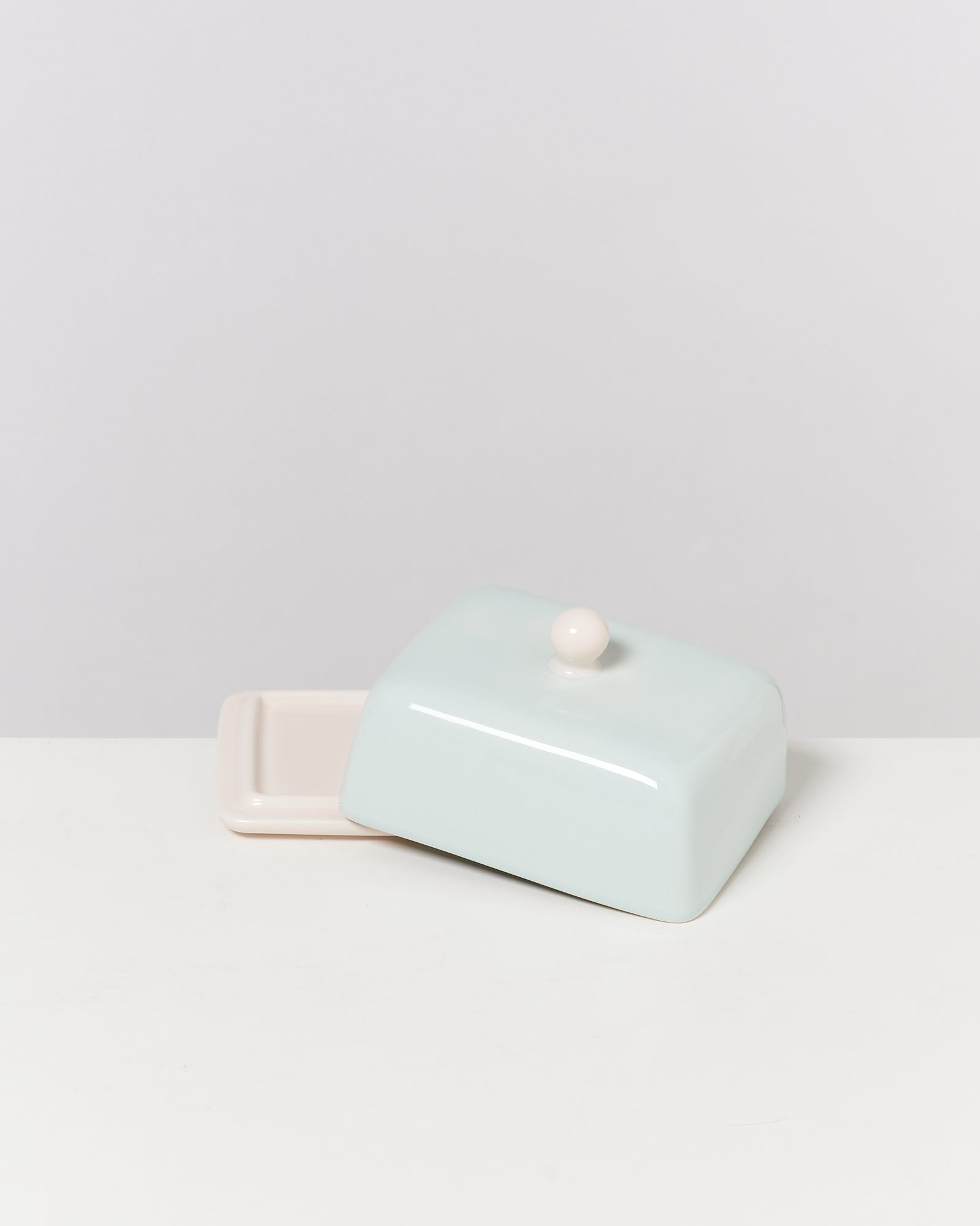 Hellblaue Butterdose mit hellem Knauf, teilweise vom Teller abgehoben, auf weißem Hintergrund.