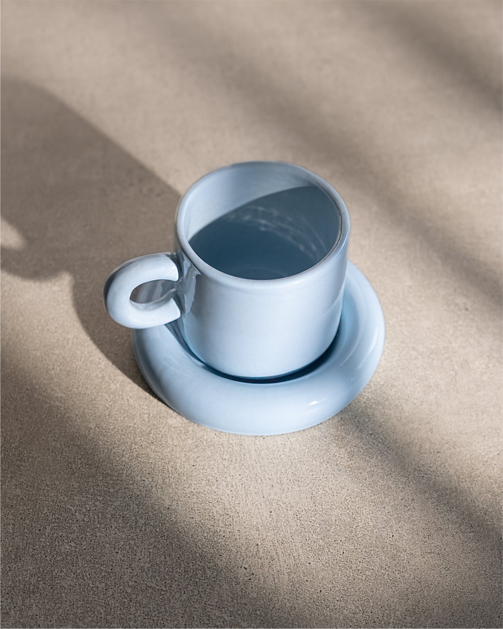 Blaue Tasse auf passendem Unterteller auf einer Betonoberfläche, im Sonnenlicht mit Schatten.
