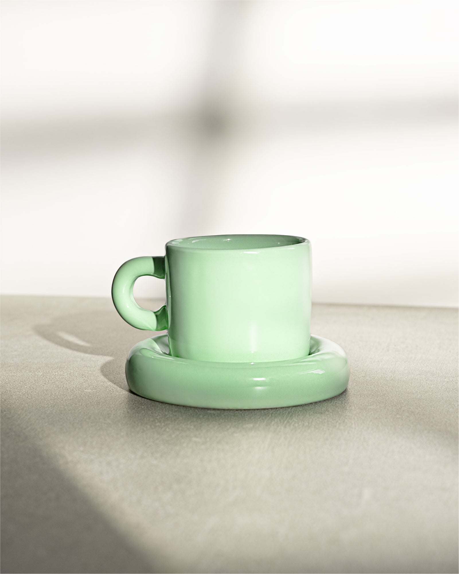 Grüne Tasse mit Untertasse auf einem neutralen Untergrund, im Sonnenlicht fotografiert.