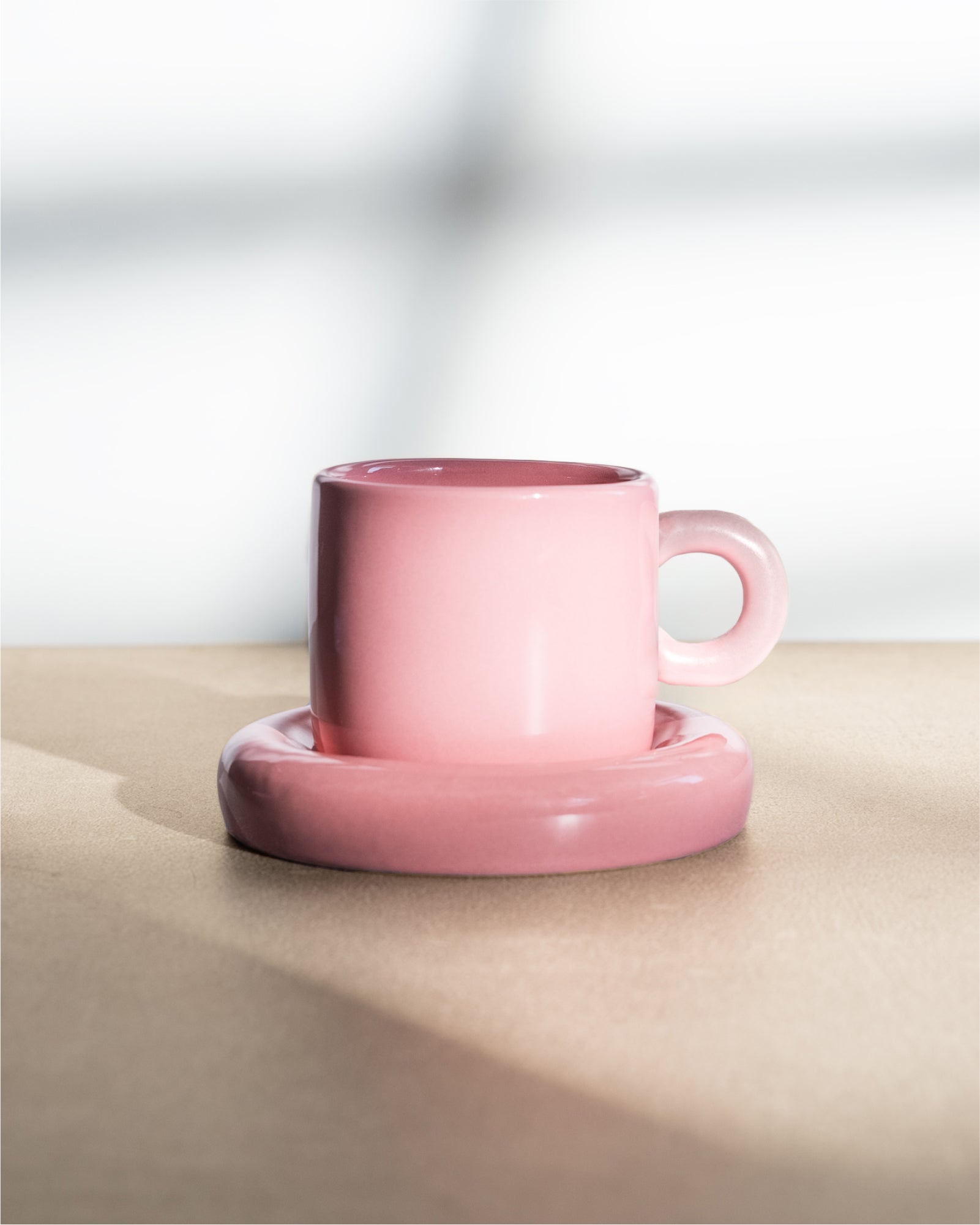 Rosa Kaffeetasse mit Untertasse auf einem Tisch vor einem unscharfen, hellen Hintergrund.