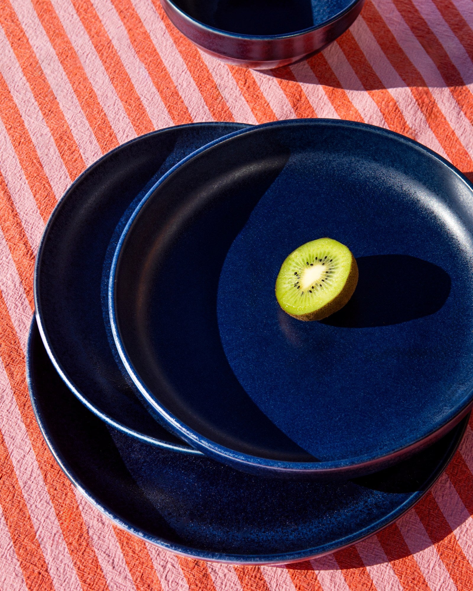 Drei blaue Teller mit einer halben Kiwi obenauf, auf einem gestreiften, rot-pinken Tischtuch.