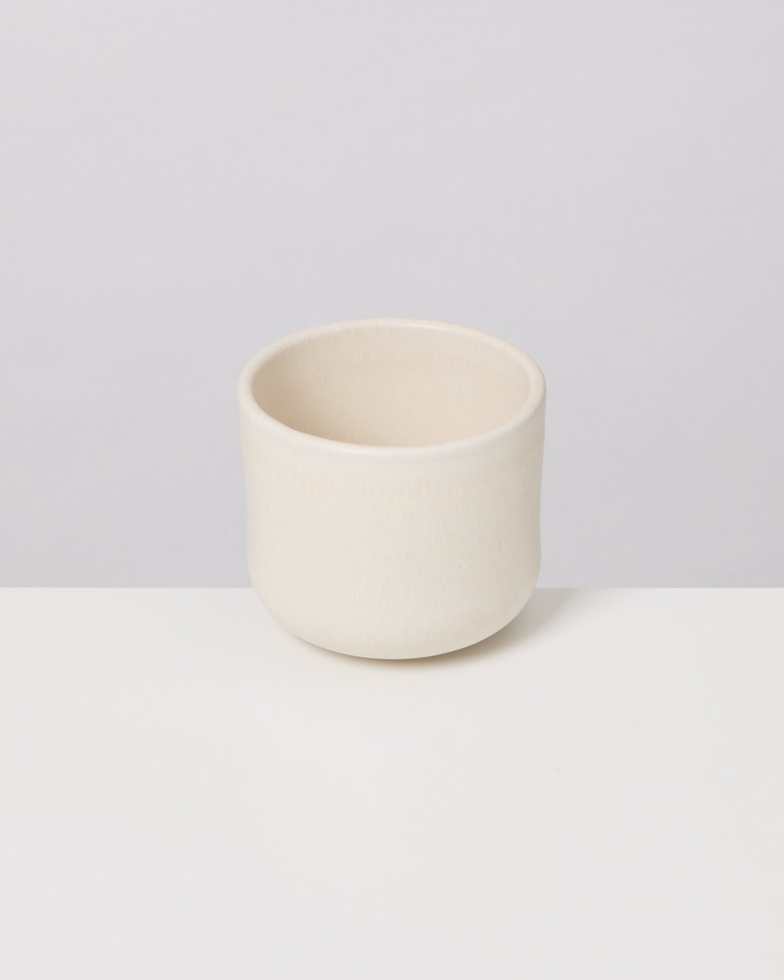 Ein cremefarbener Keramikbecher steht auf einer weißen Oberfläche vor einem grauen Hintergrund.