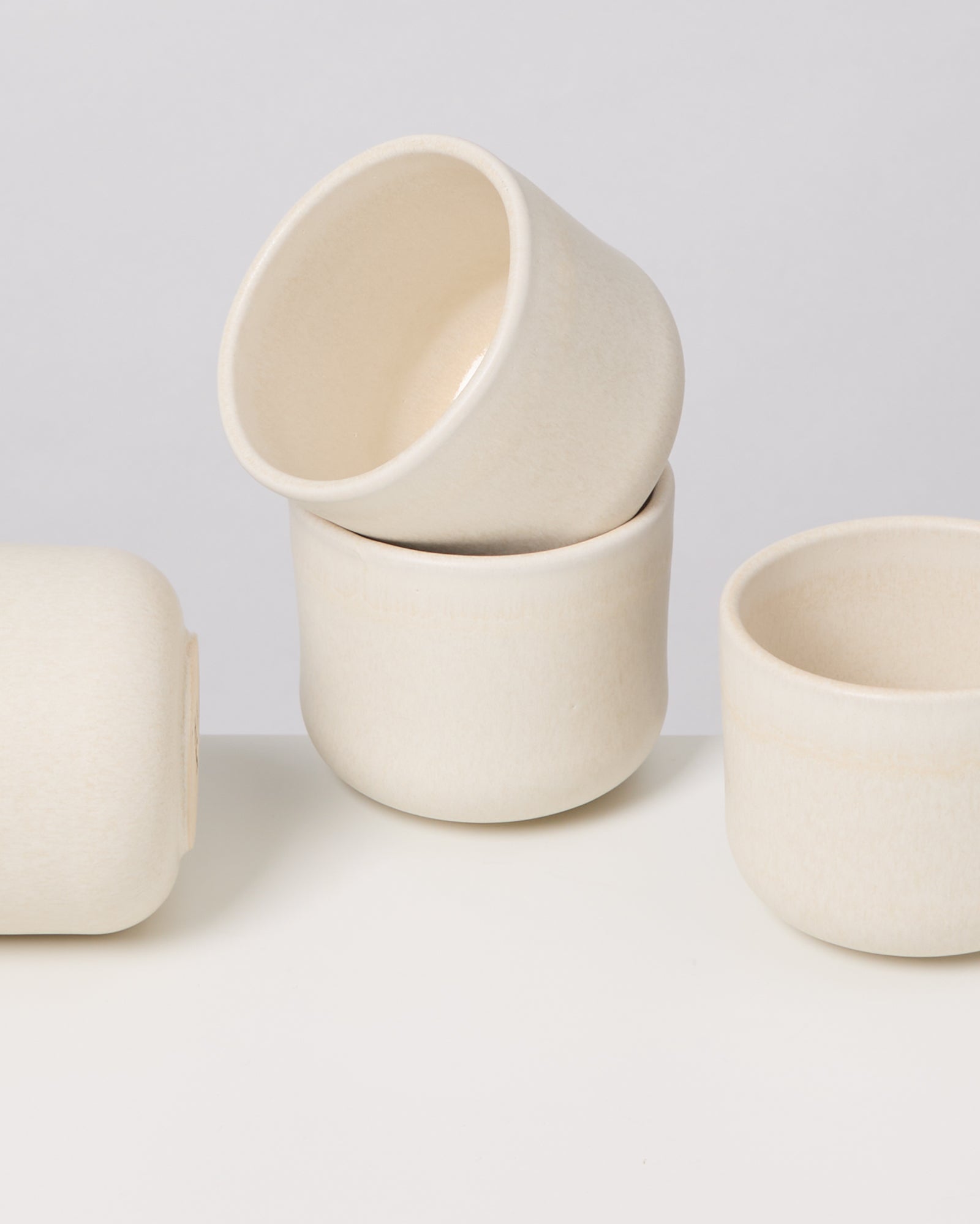 Vier weiße, minimalistische Keramiktassen auf einer weißen Oberfläche, eine kopfüber gestapelt.