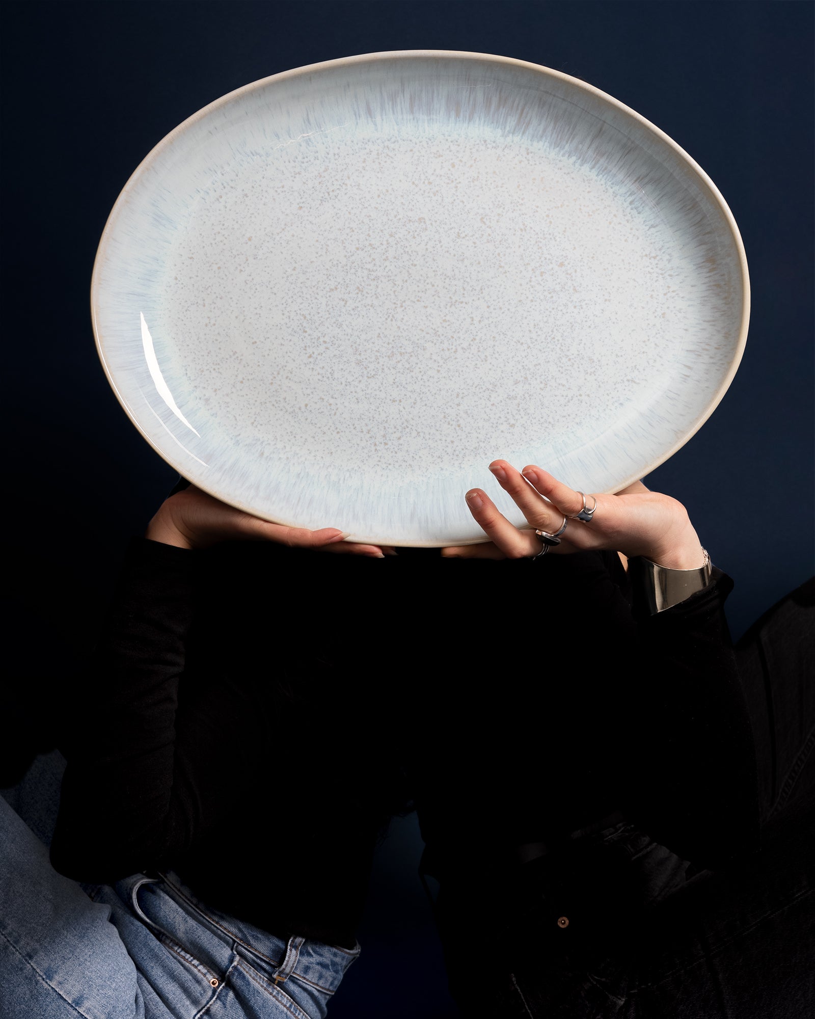 Person hält einen großen, weißen ovalen Teller vor dem Gesicht, vor dunkelblauem Hintergrund.