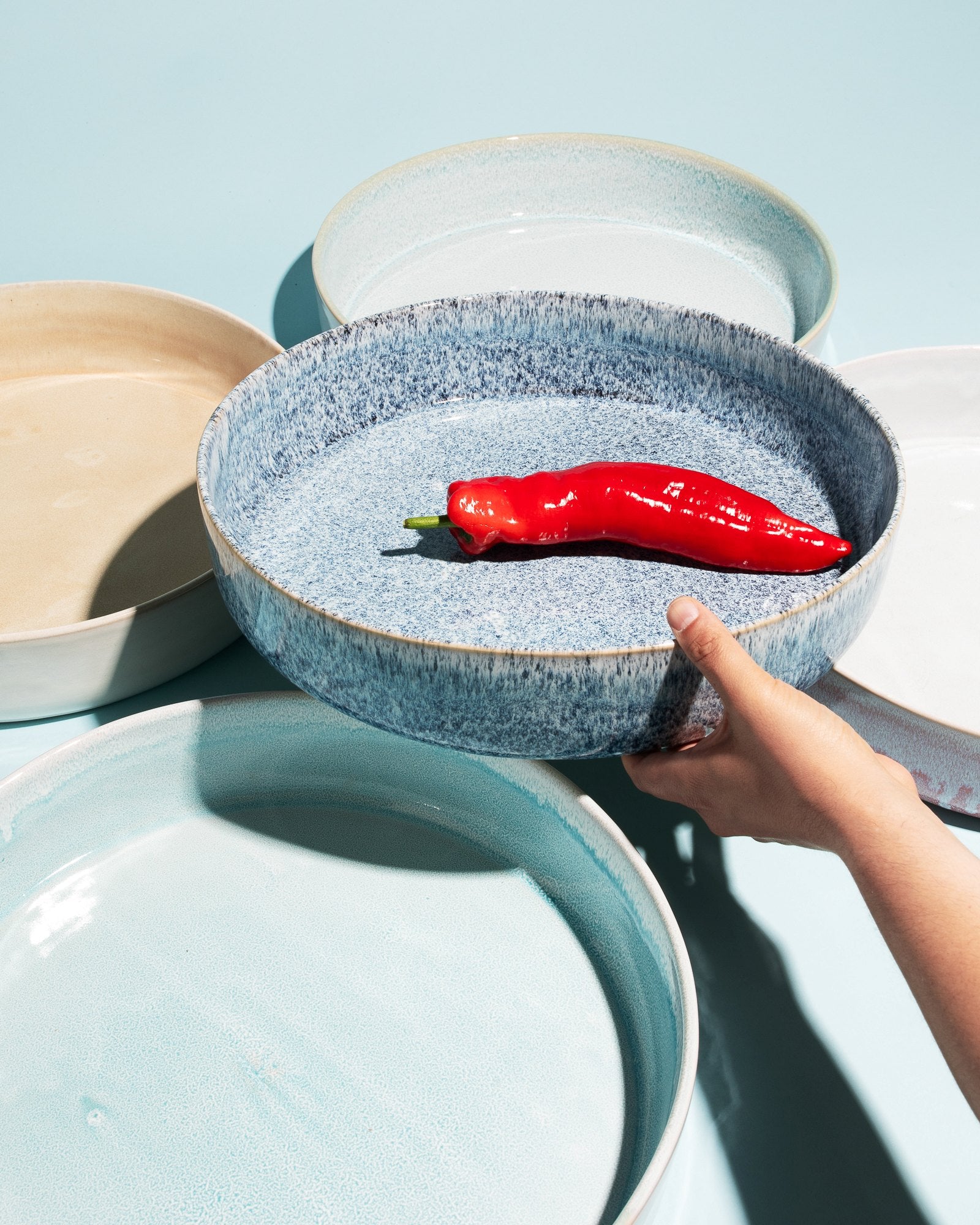 Eine Hand hält eine blaue Schale mit einer roten Paprika darauf, umgeben von anderen Schalen auf hellblauem Hintergrund.