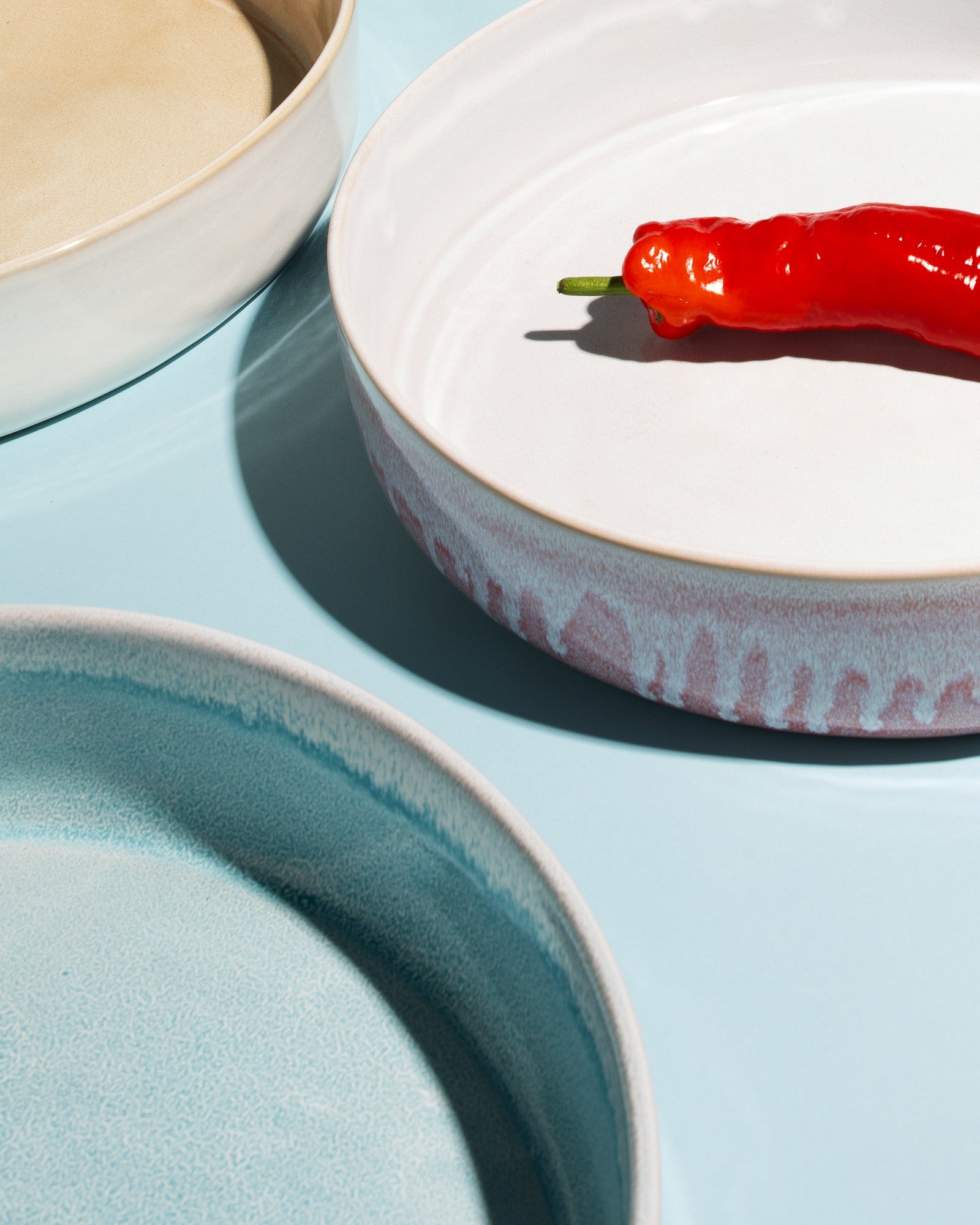 Drei runde, verschiedenfarbige Keramikteller auf einer blauen Oberfläche, einer mit einer roten Chilischote.