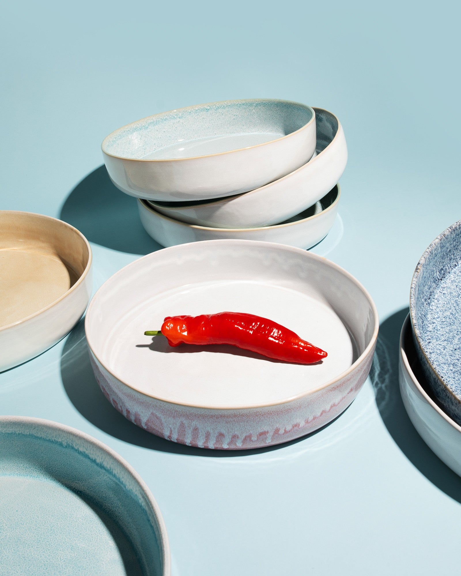 Rote Chilischote auf einem weißen Teller, umgeben von mehreren gestapelten, farblich abgestimmten Tellern auf blauem Hintergrund.