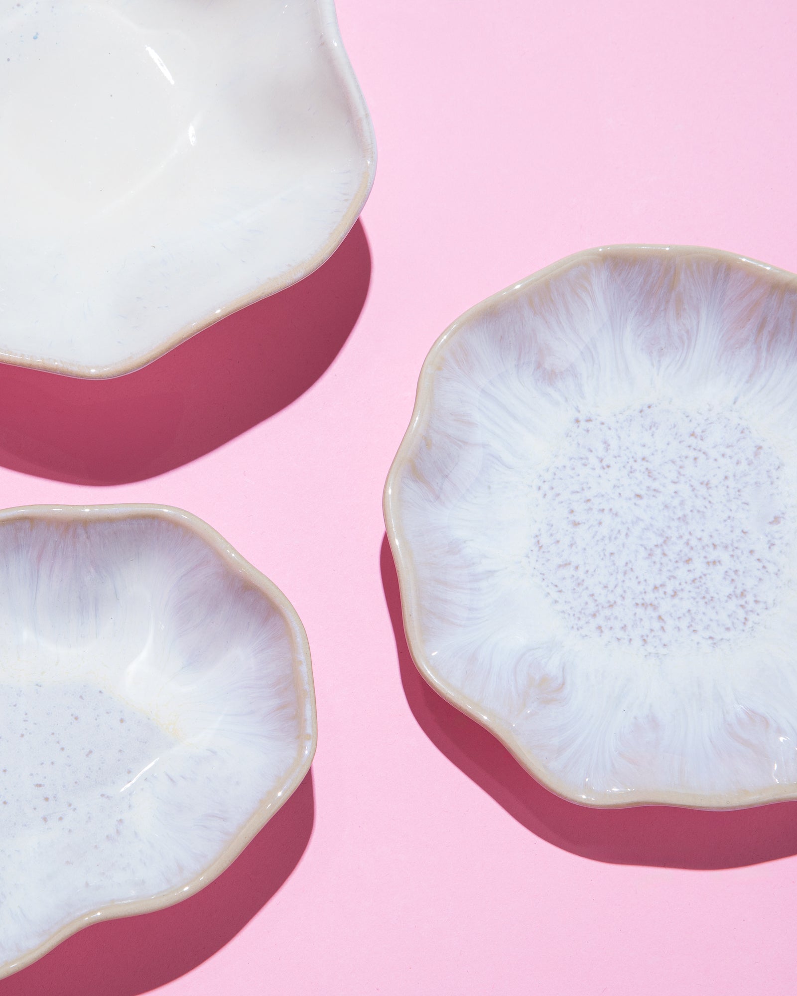 Drei weiße, blumenförmige Teller mit gewelltem Rand auf rosa Hintergrund.