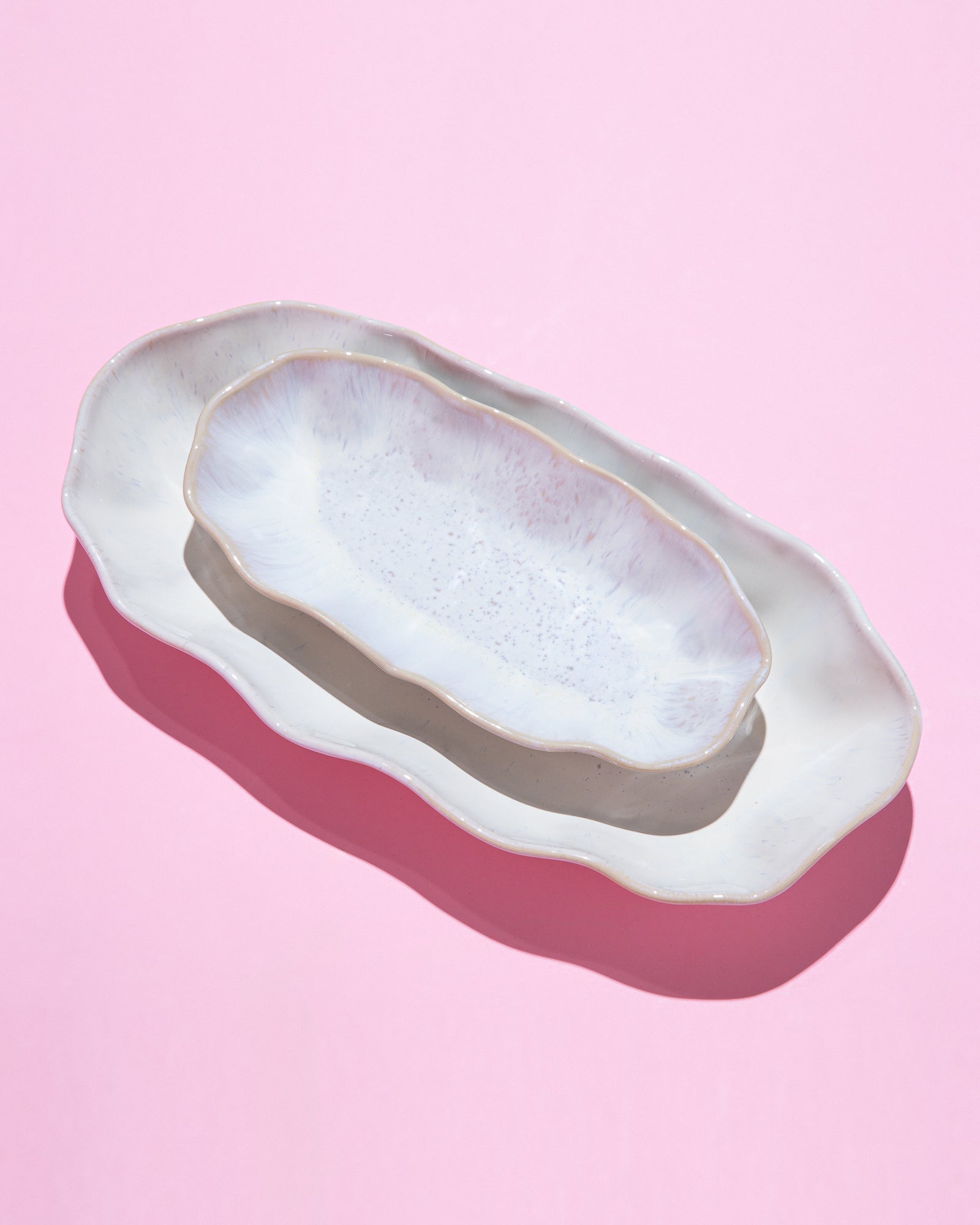Zwei längliche, gewellte, weiße Keramikplatten auf einem rosa Hintergrund, im Licht fotografiert.