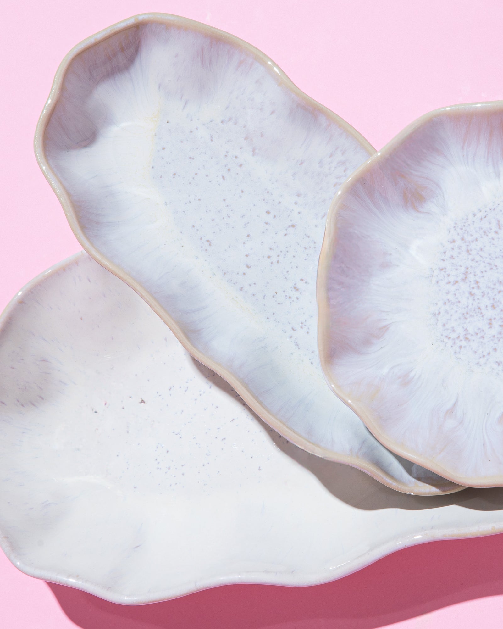 Drei weiße, unregelmäßig geformte Keramikteller auf rosa Hintergrund.