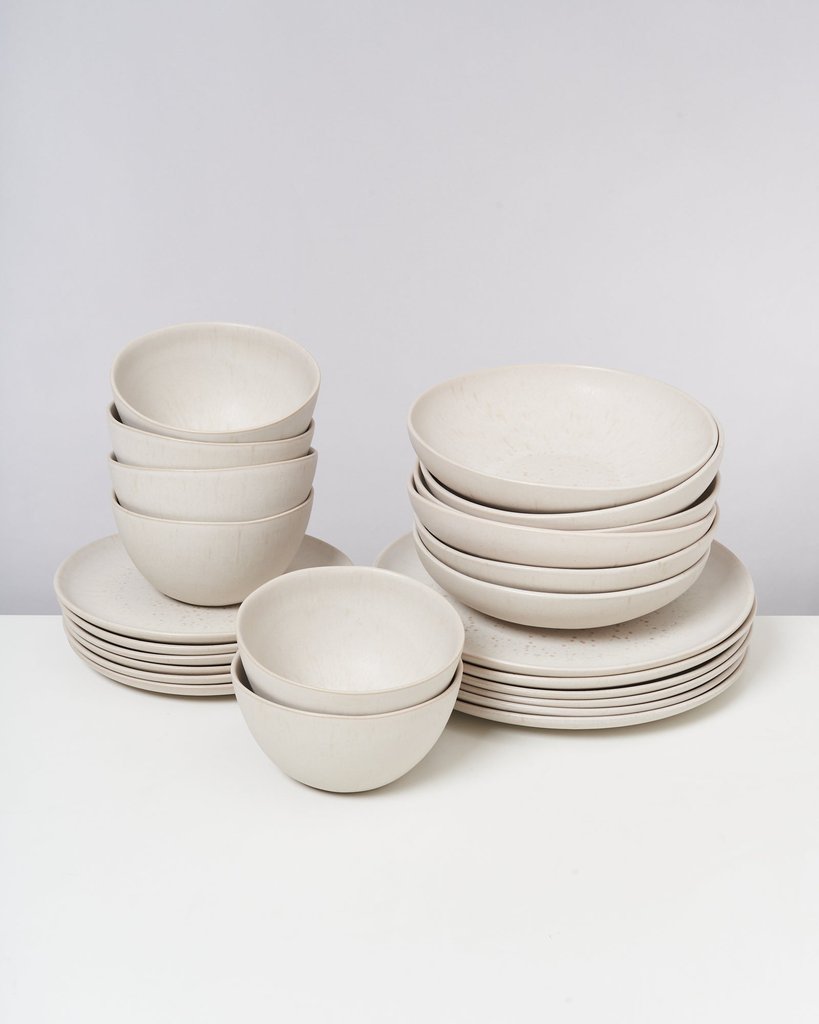 Ein Stapel aus cremefarbenen Schüsseln und Tellern auf einem weißen Tisch vor einem grauen Hintergrund.