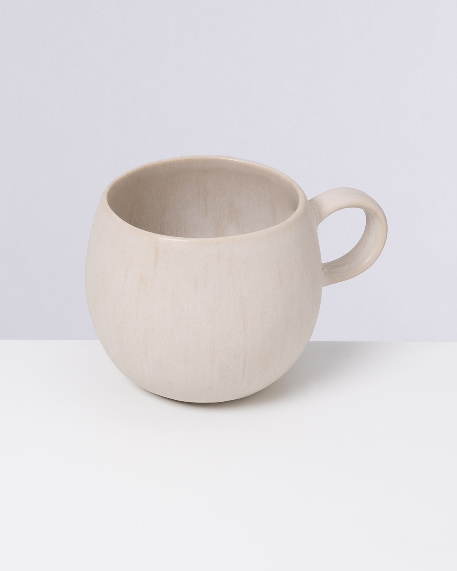 Cremefarbene Keramiktasse mit rundem Körper und Henkel, auf weißem Hintergrund.