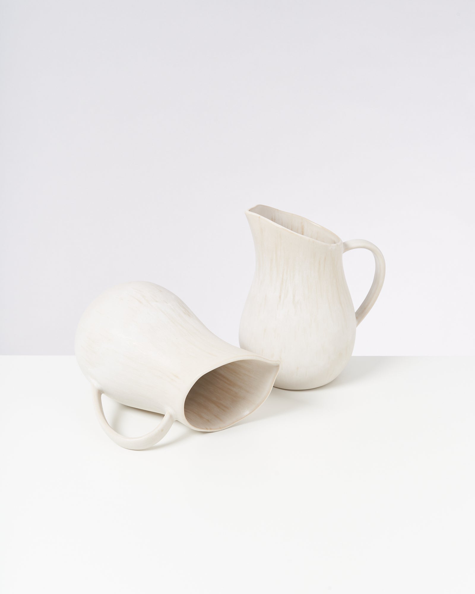 Zwei weiße Krüge, einer liegend und einer stehend, auf einem weißen Tisch vor neutralem Hintergrund.