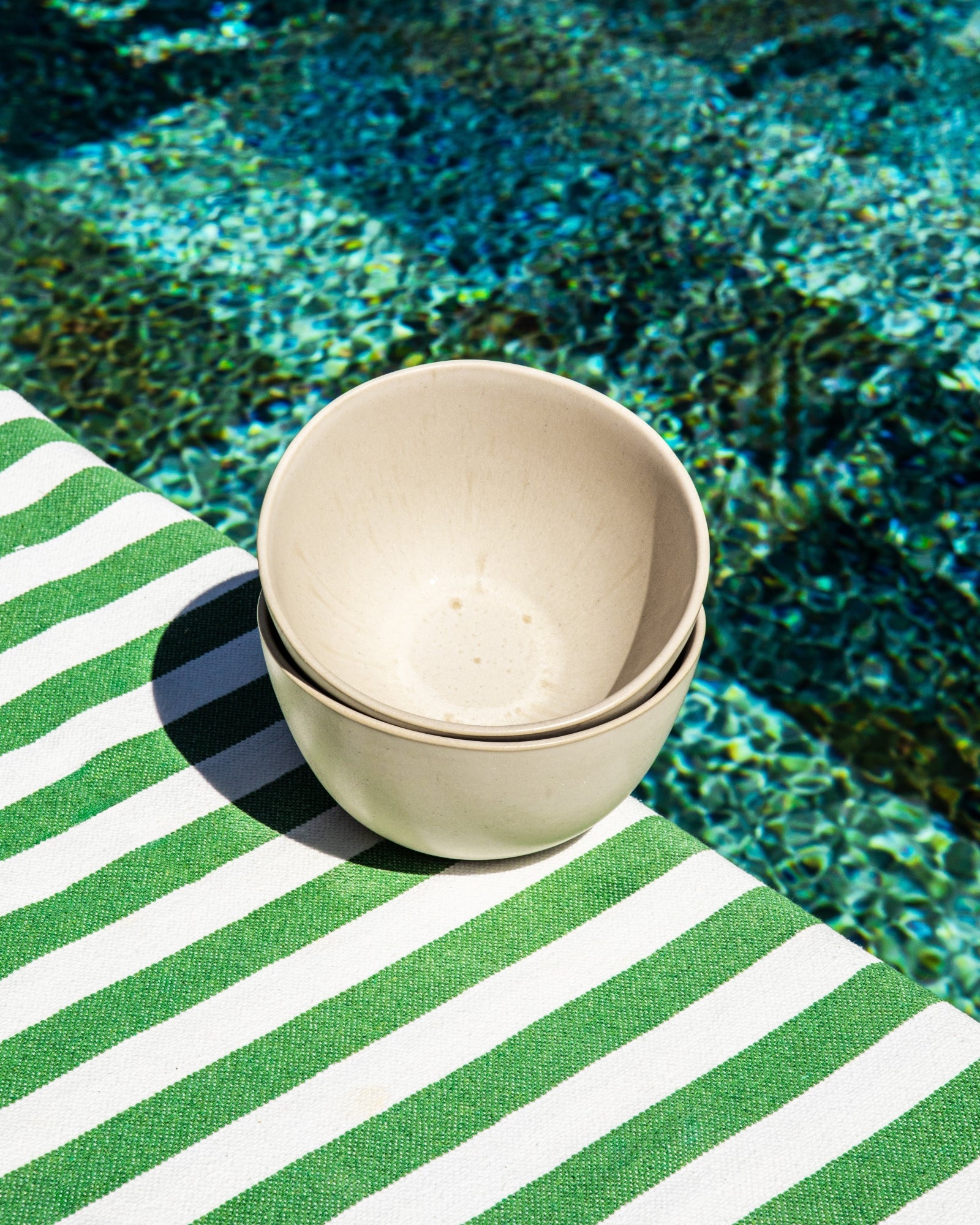 Zwei helle Schalen auf einem grün-weiß gestreiften Tuch, nahe einem Pool mit blauem Wasser.