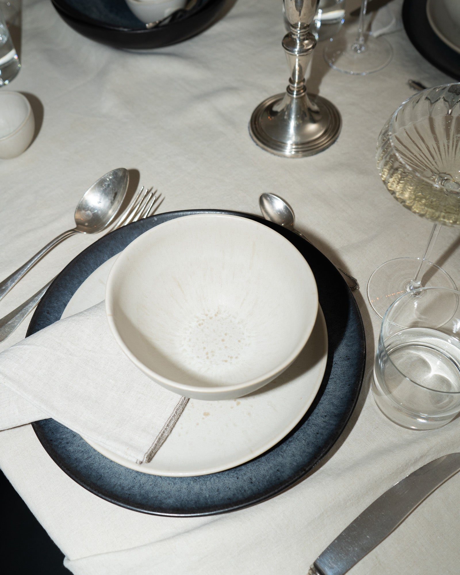 Elegantes Gedeck mit weißem Geschirr, Gläsern und Besteck auf einem weißen Tisch mit Stoffserviette und Kerzenhalter.