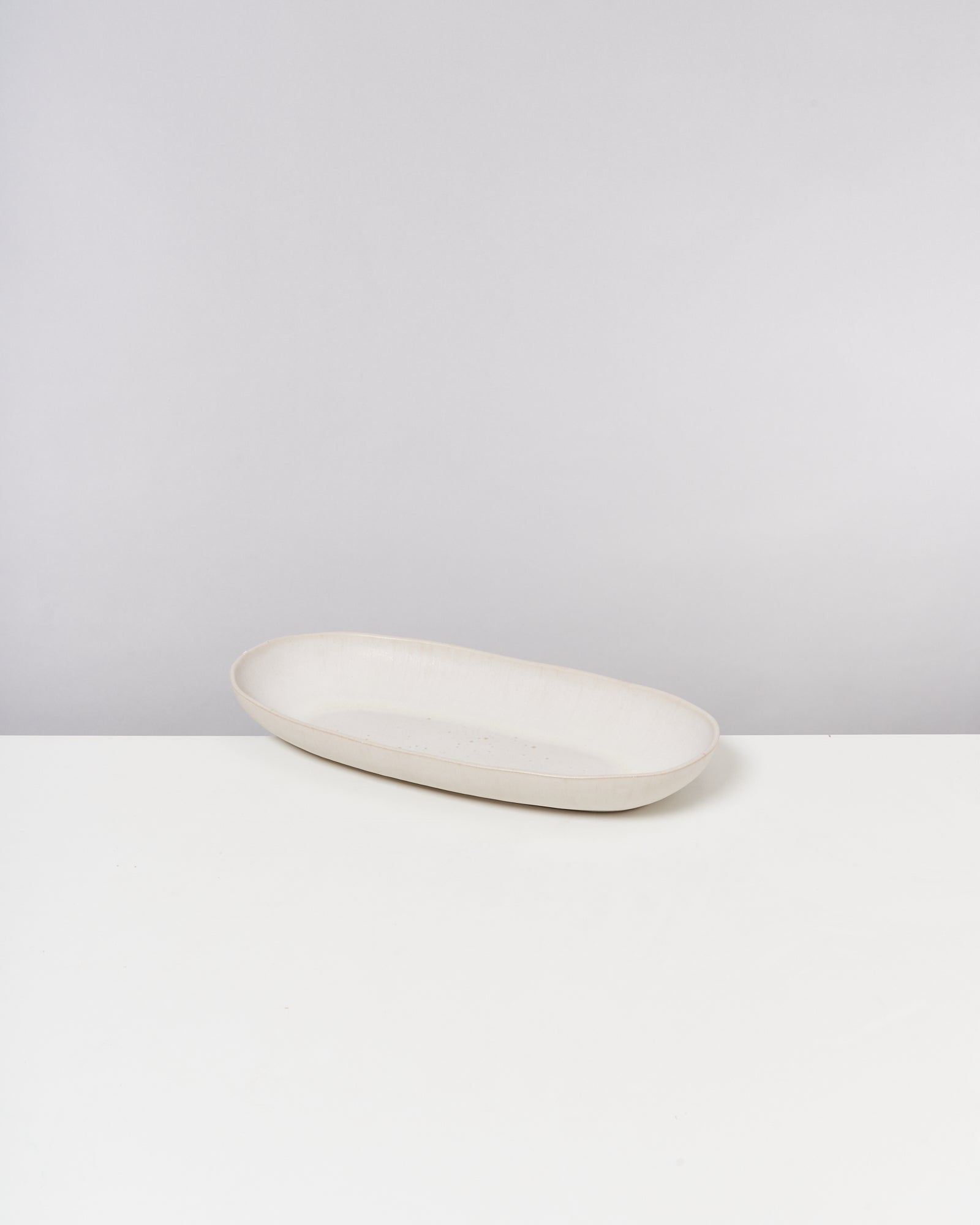 Ovale, cremefarbene Keramikschale auf weißem Tisch vor grauem Hintergrund.