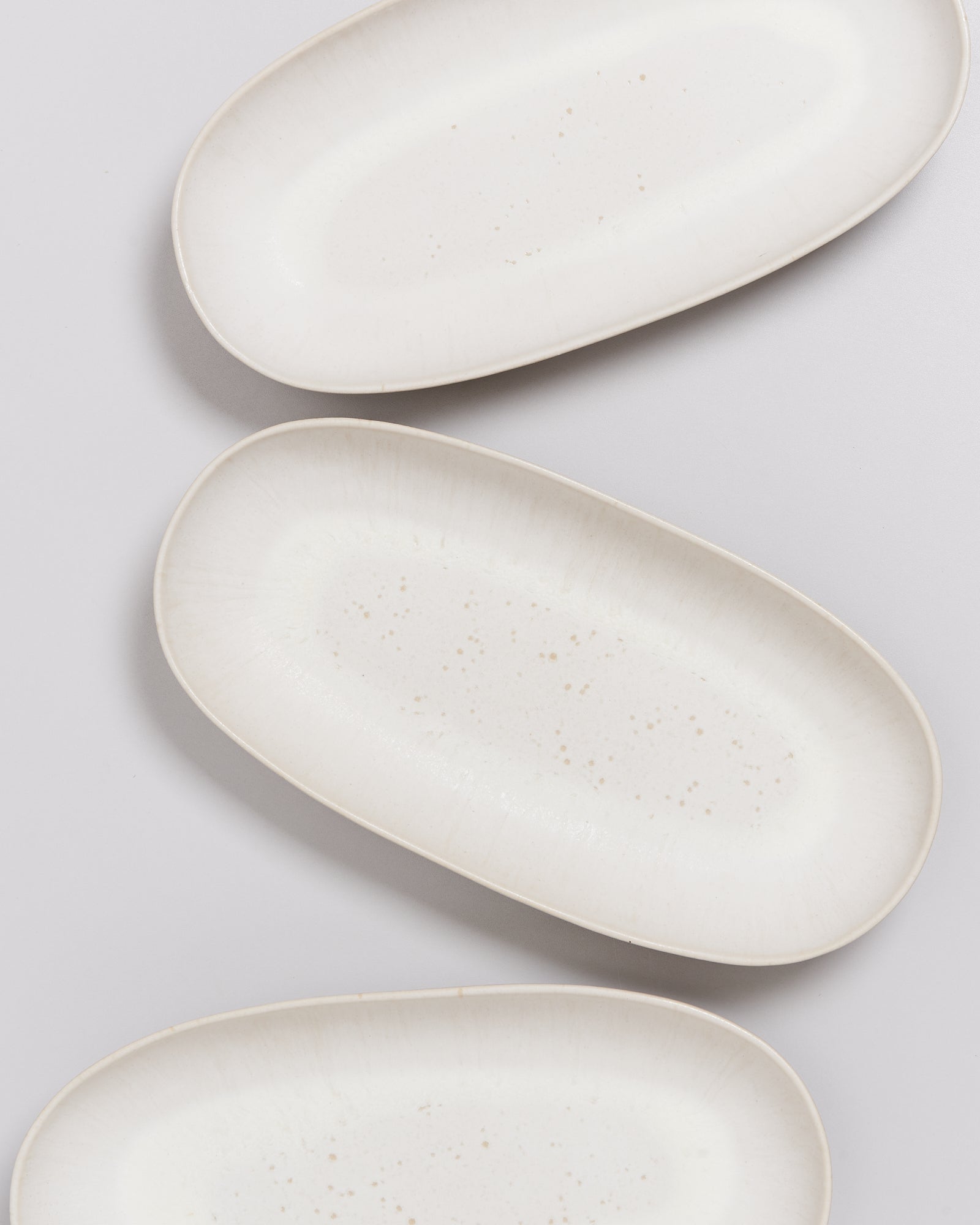 Drei weiße, ovale Teller mit dezent gesprenkeltem Muster auf einer hellen Oberfläche angeordnet.
