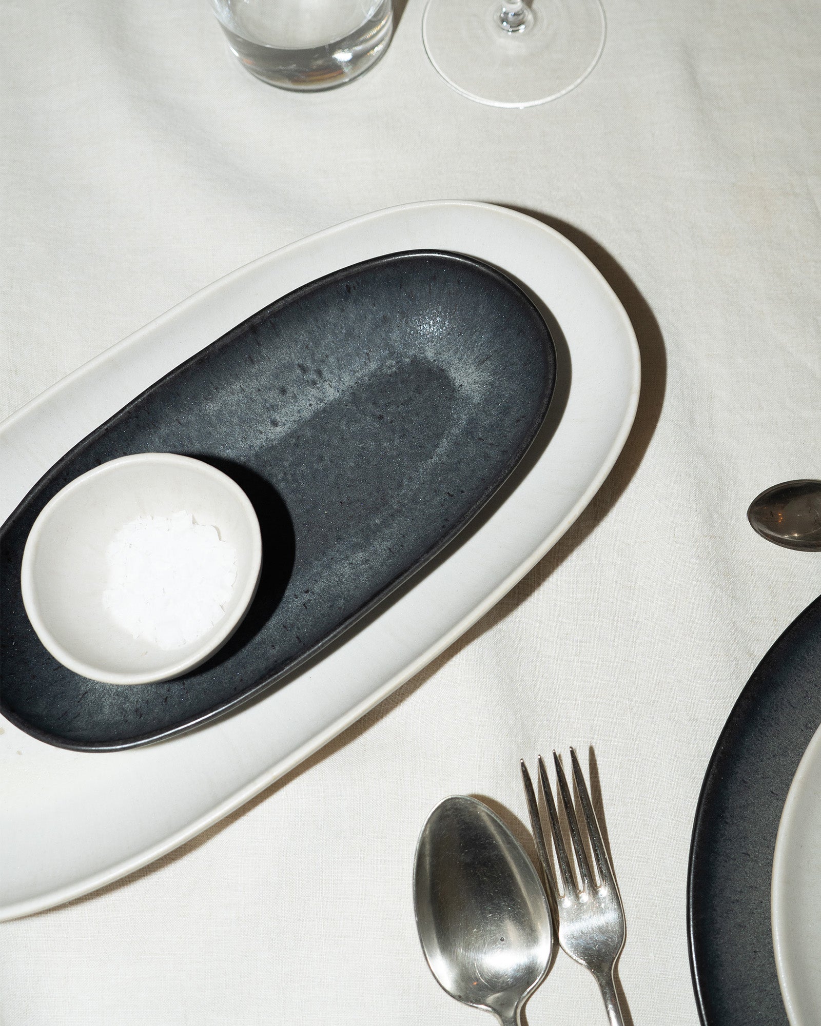 Weiß gedeckter Tisch mit schwarzem Teller, kleiner Schale mit Salz, Besteck und Glas.