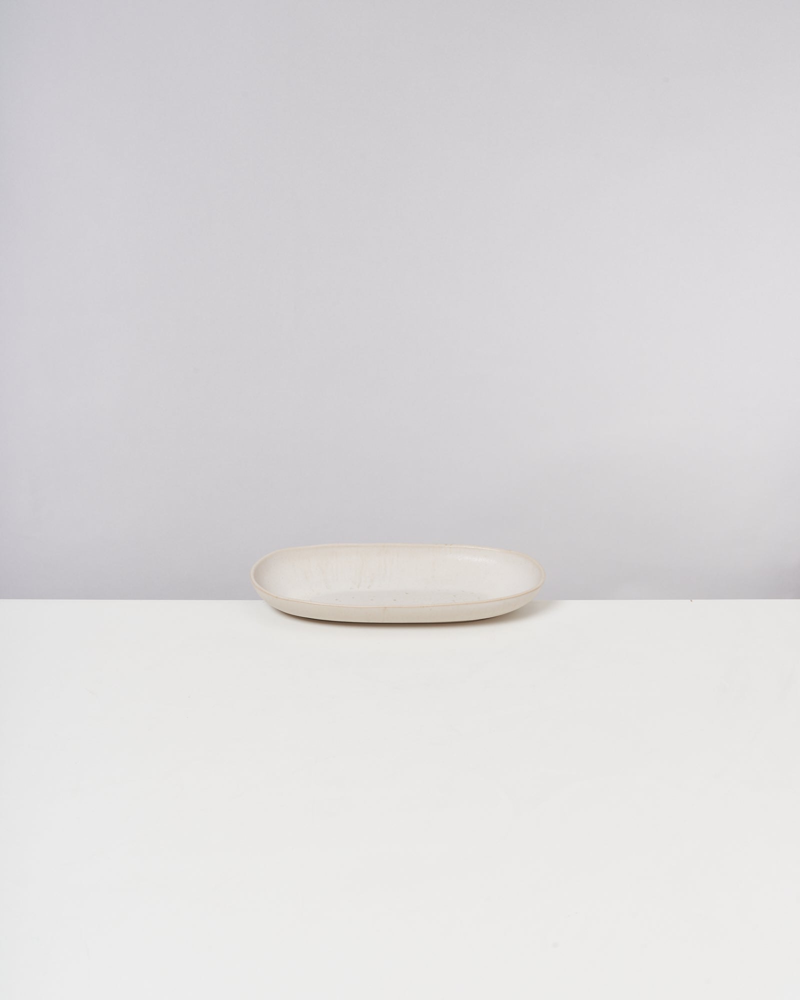 Ein ovaler, cremefarbener Keramikteller steht auf einem weißen Tisch vor einem neutralen Hintergrund.