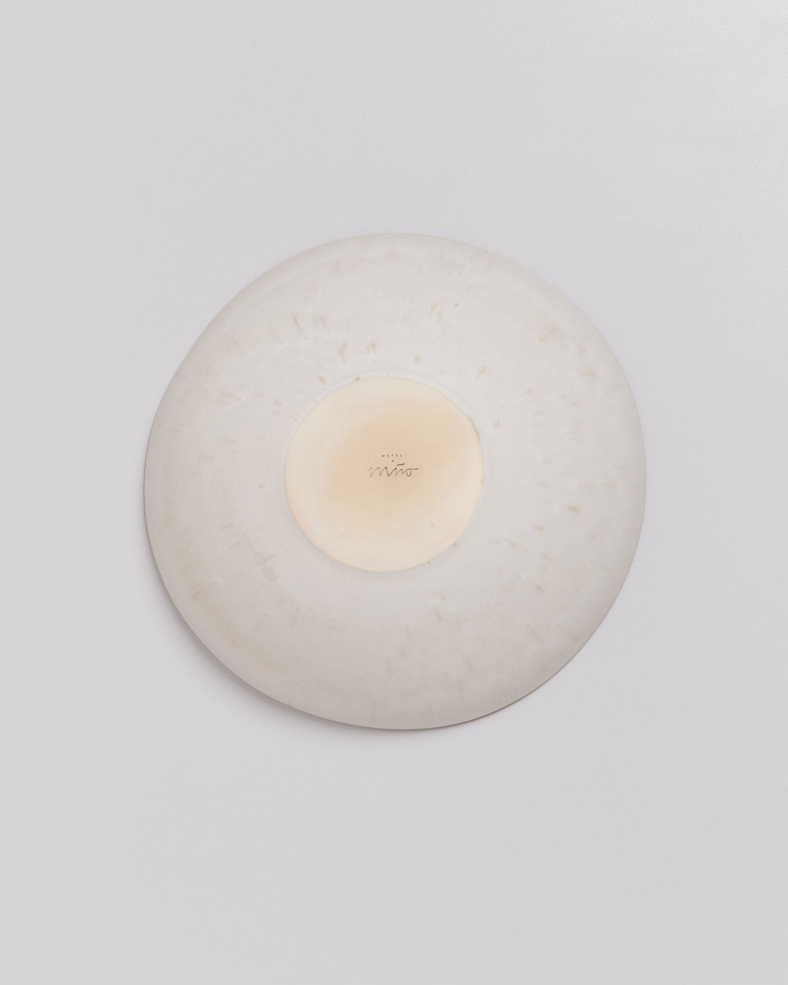 Rückseite eines weißen Keramiktellers mit mittiegem Markensiegel und Unterschrift auf grauem Hintergrund.