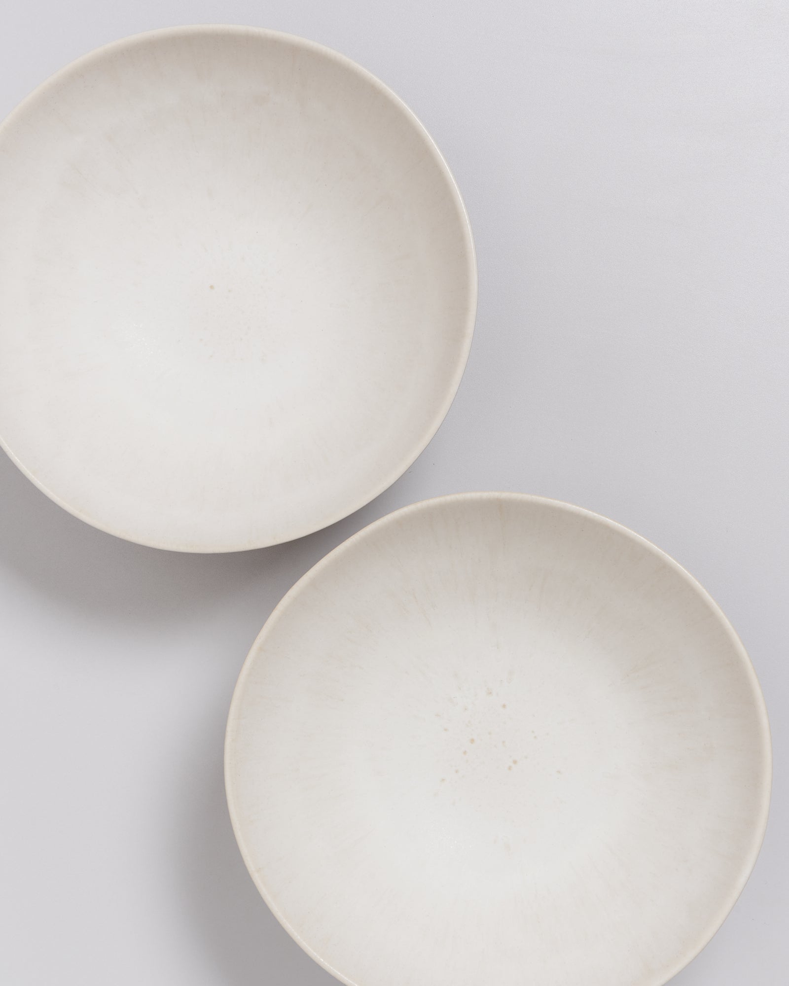 Zwei weiße, runde Teller mit leichtem Muster liegen auf einer hellgrauen Oberfläche.