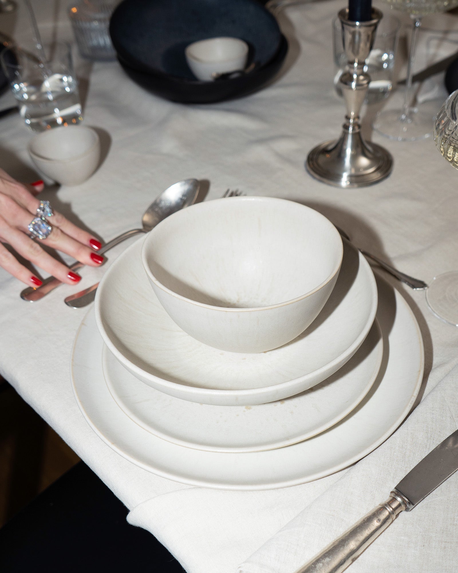 Ein gedeckter Tisch mit weißen Tellern und Schüsseln, Besteck und einem Glas auf einer Tischdecke, eine Hand mit rotem Nagellack.
