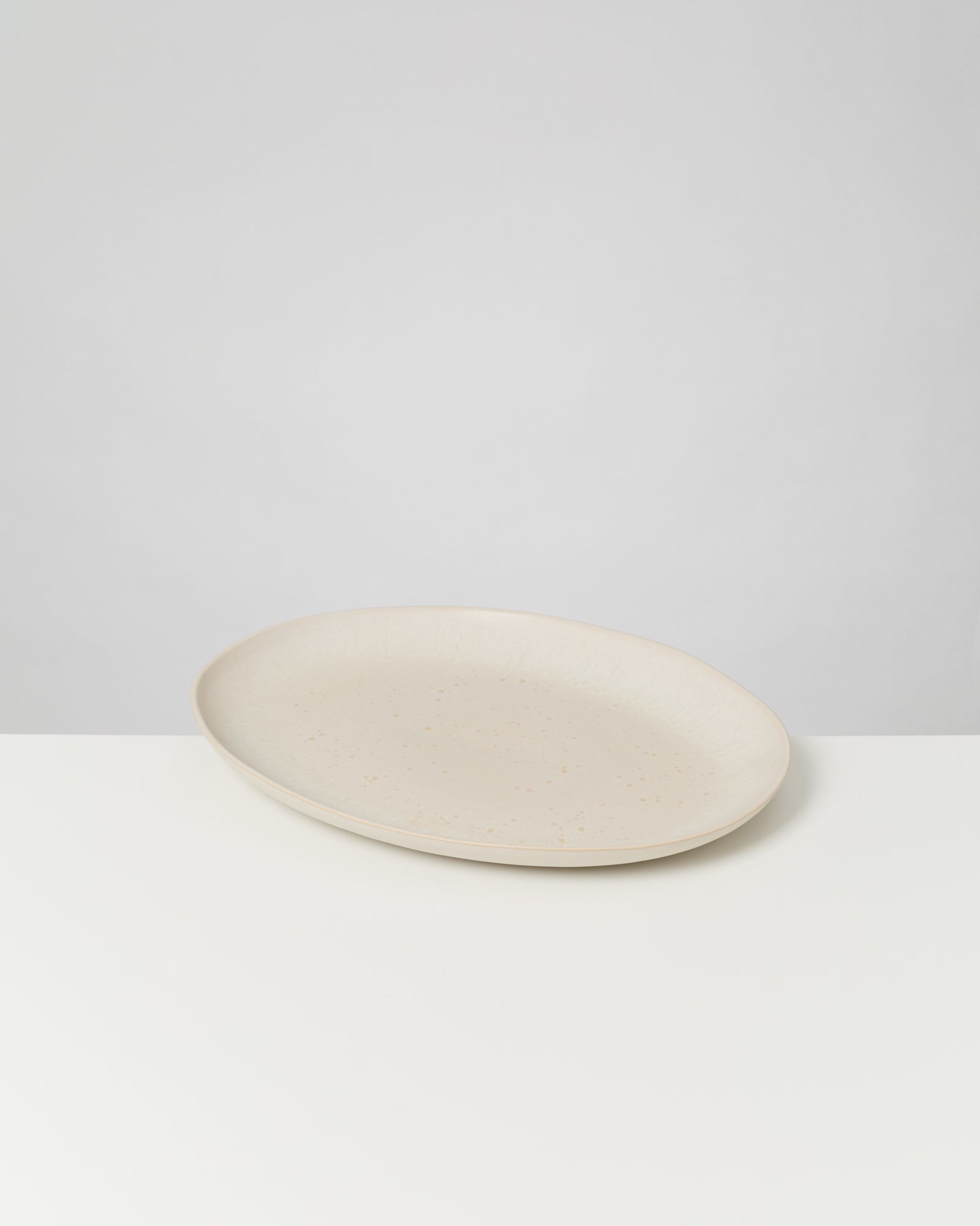 Flacher, beigefarbener Keramikteller auf einem weißen Tisch vor einem grauen Hintergrund.