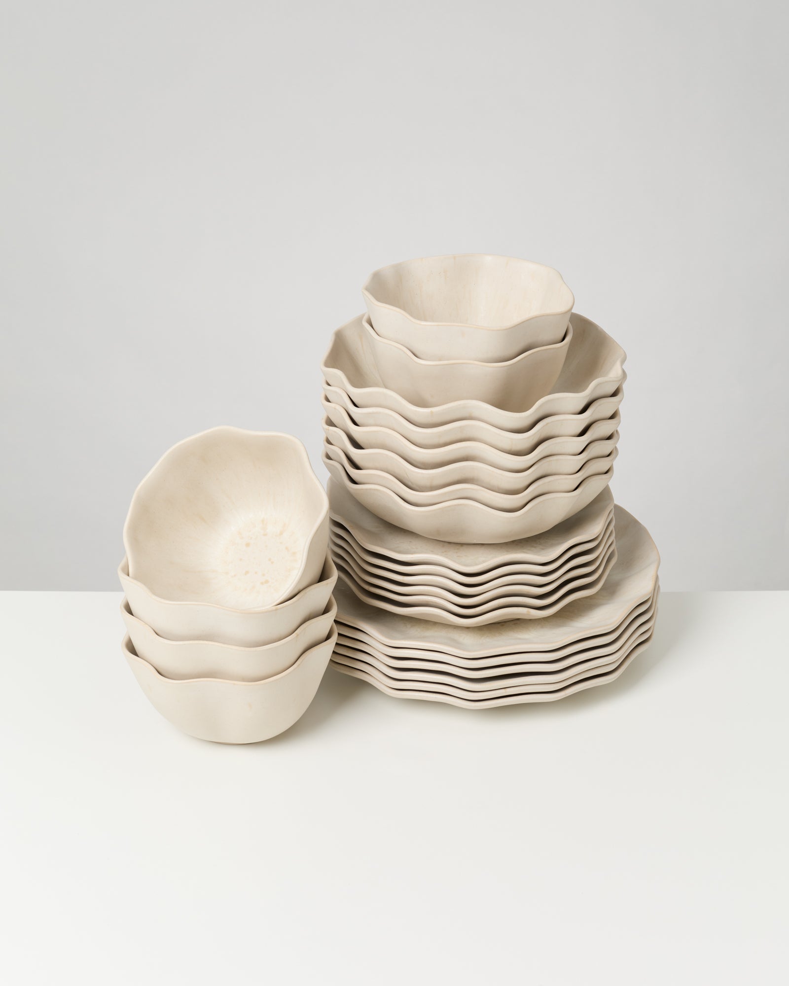Stapel von cremefarbenen, wellenförmigen Keramikschalen und -tellern auf einem weißen Untergrund.