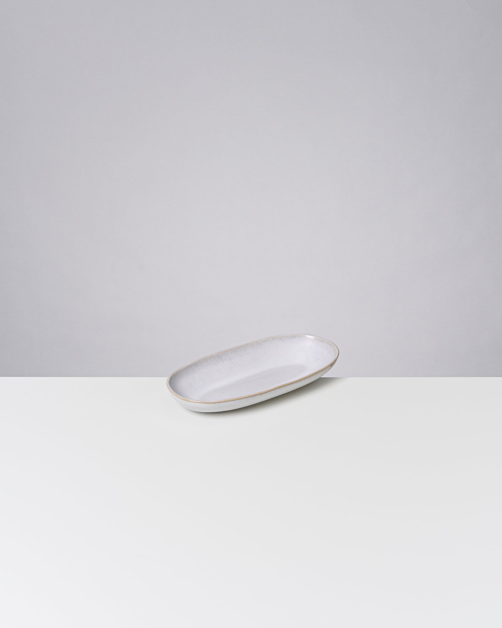 Ein ovaler, grauer Keramikteller liegt auf einer weißen Oberfläche vor einem grauen Hintergrund.
