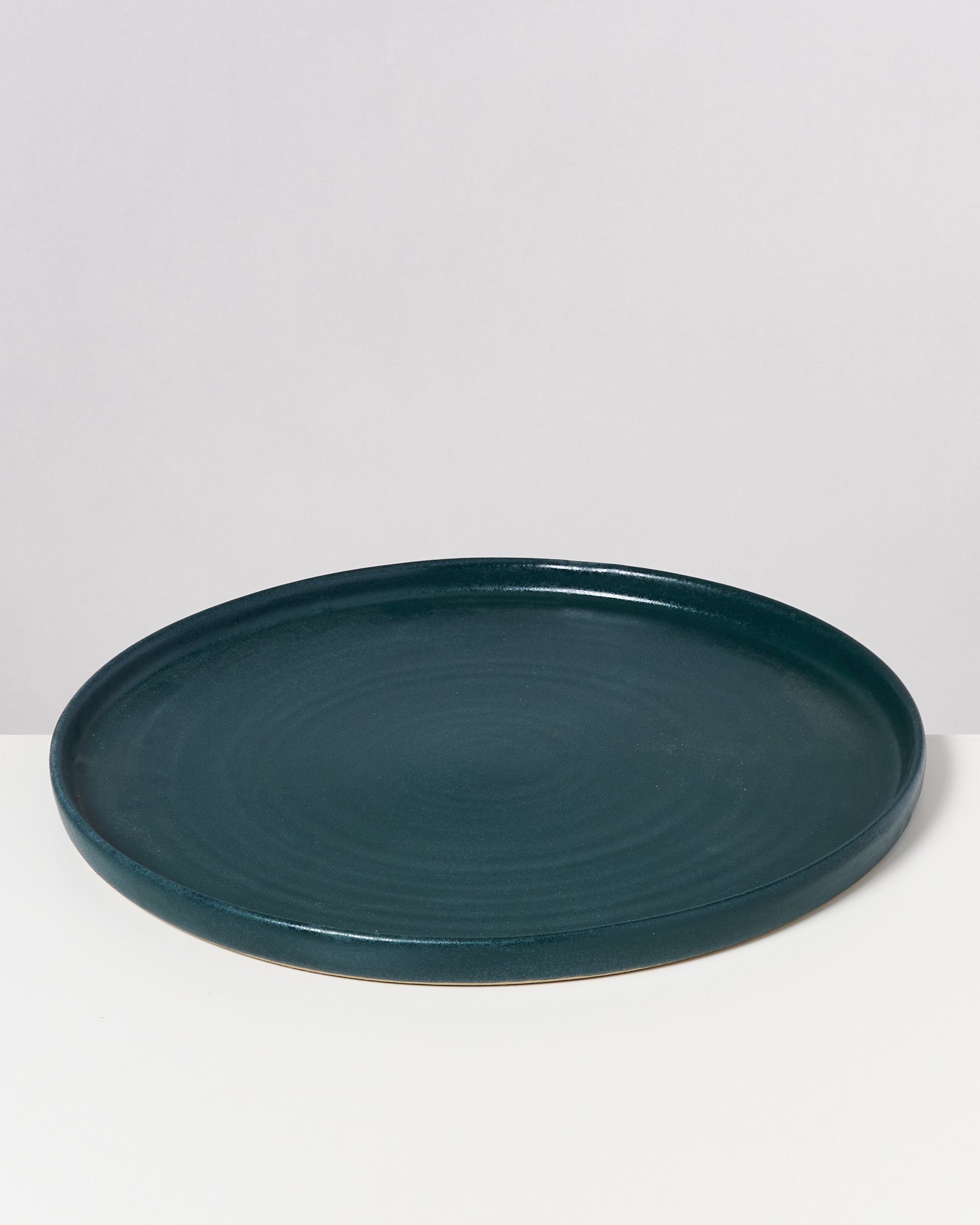 Dunkelgrüner, runder Teller auf einer weißen Oberfläche vor einem hellgrauen Hintergrund.