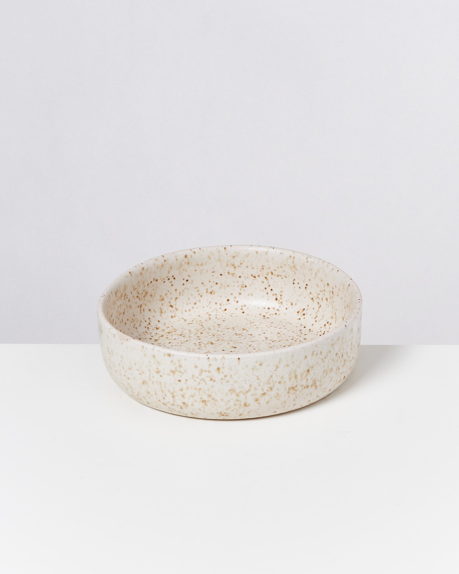 Keramikschale in Beige mit braunen Sprenkeln steht auf einem weißen Tisch vor einem hellen Hintergrund.