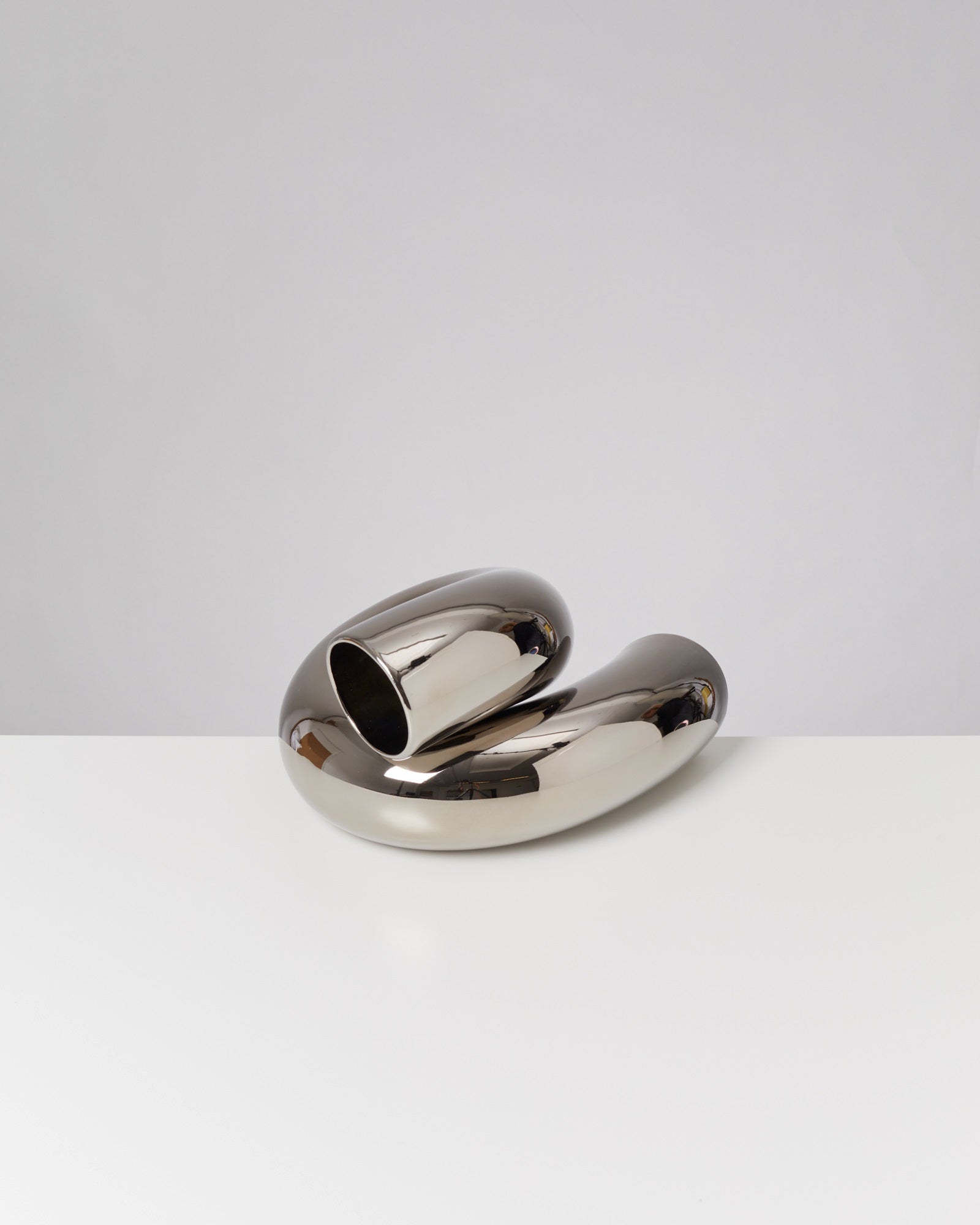 Metallische, geschwungene Skulptur auf weißem Tisch, vor grauem Hintergrund.