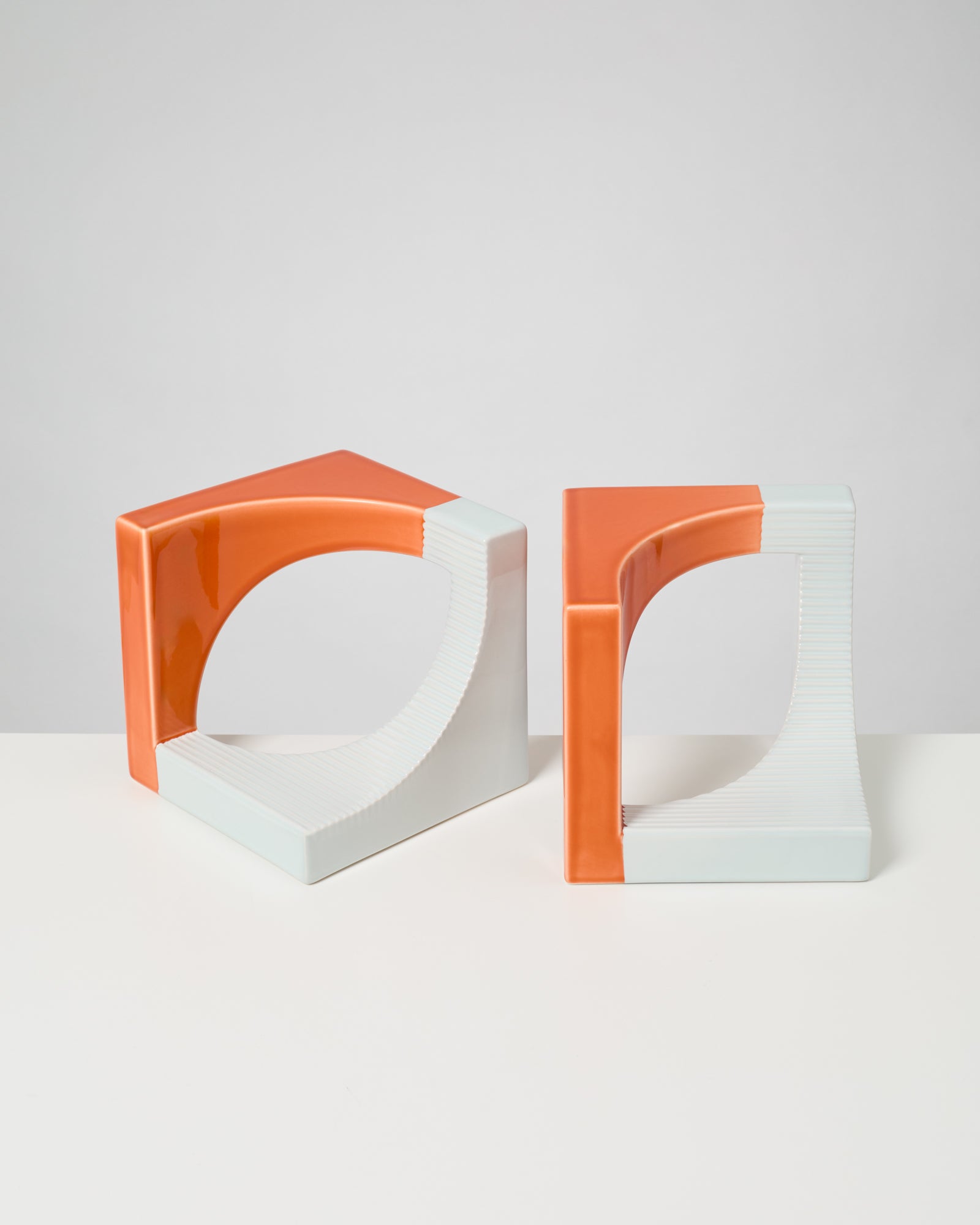 Zwei moderne, abstrakte orange-weiße Skulpturen mit offenen Kreisformen auf einem weißen Tisch vor grauem Hintergrund.