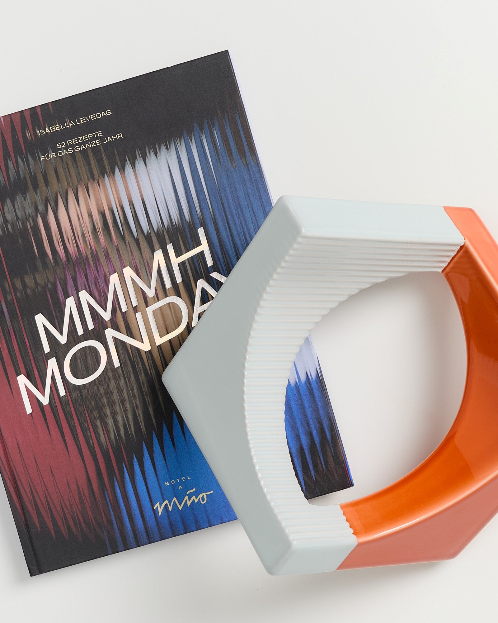 Buch mit dem Titel "MMMH MONDAY" neben einem abstrakten, orange-weißen Keramikobjekt auf einem Tisch.