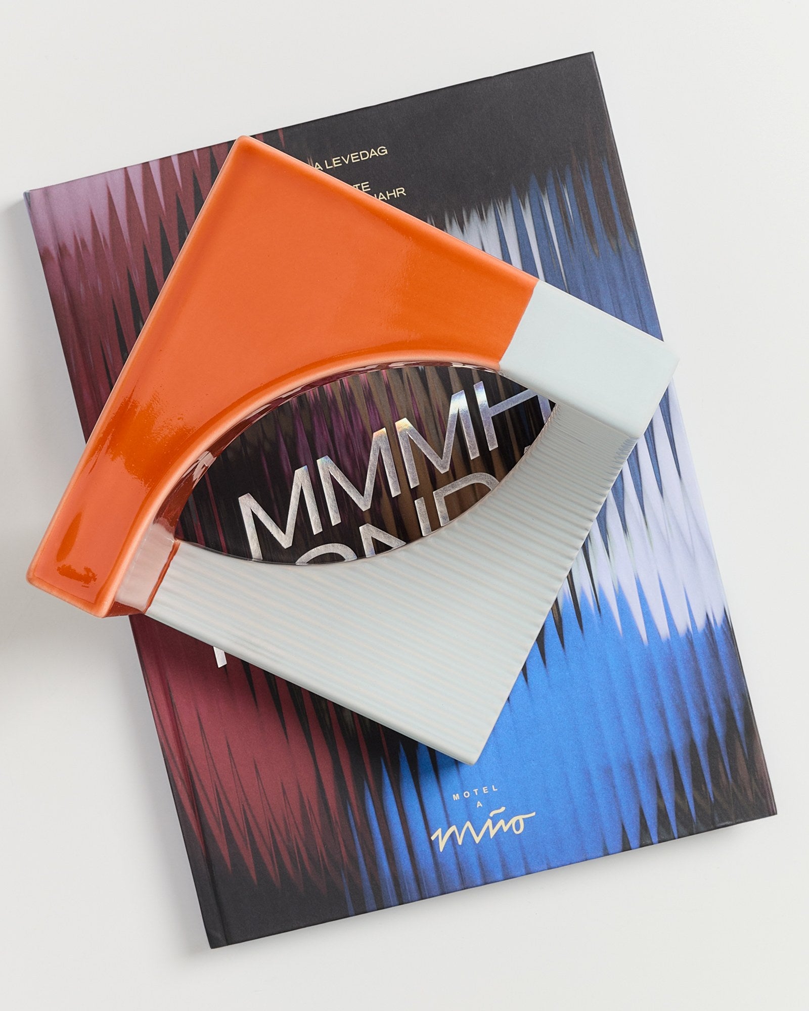 Ein orange-weißes Keramikstück liegt auf einem farbigen Buch mit der Aufschrift "MMMH" und "Motel a Miio".