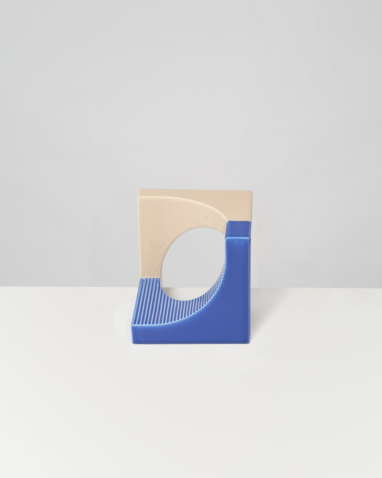 Beigefarbenes und blaues geometrisches Objekt mit kreisförmiger Öffnung auf grauem Hintergrund.