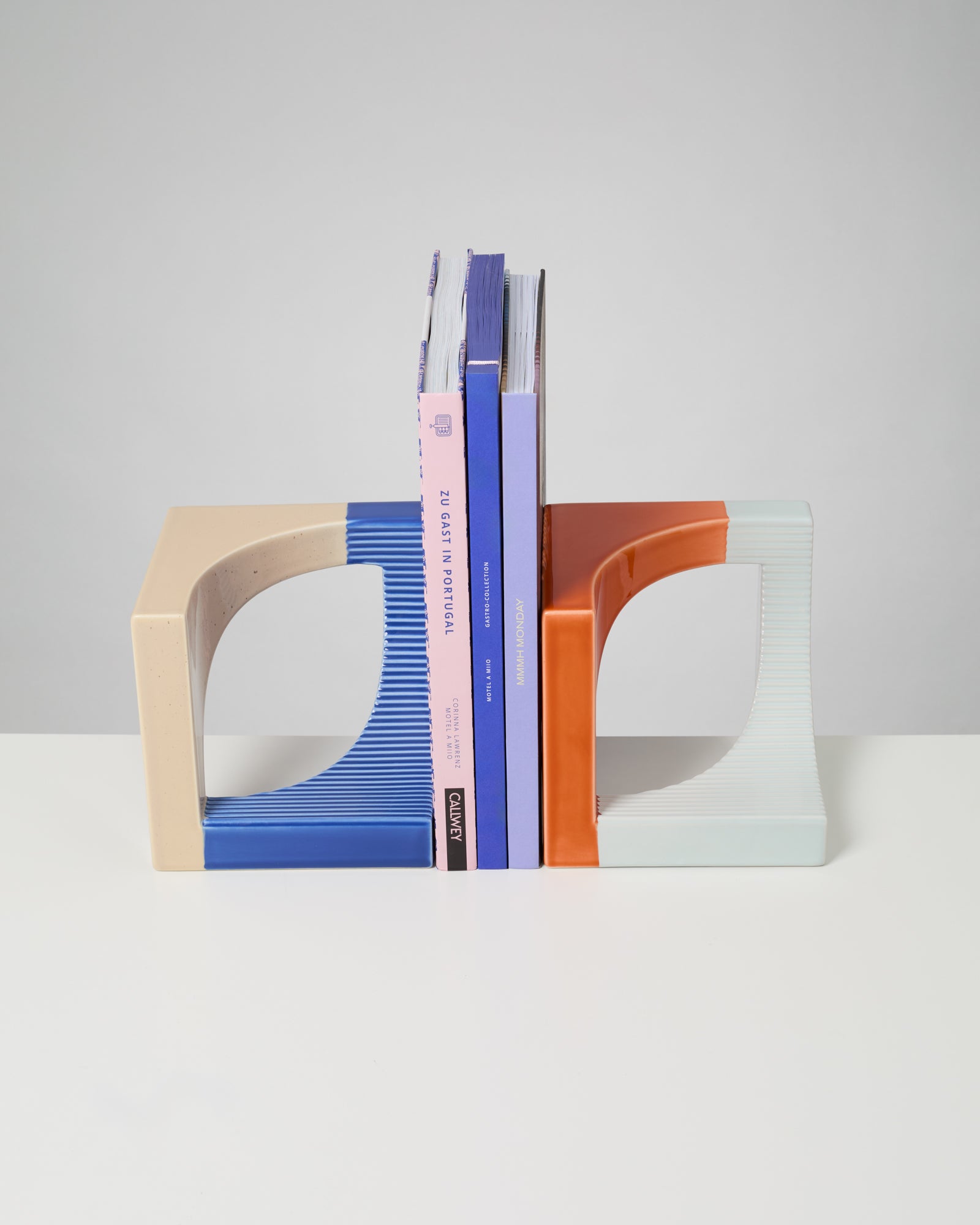 Bücher stehen zwischen zwei farbigen, gebogenen Buchstützen auf einem weißen Tisch vor grauem Hintergrund.