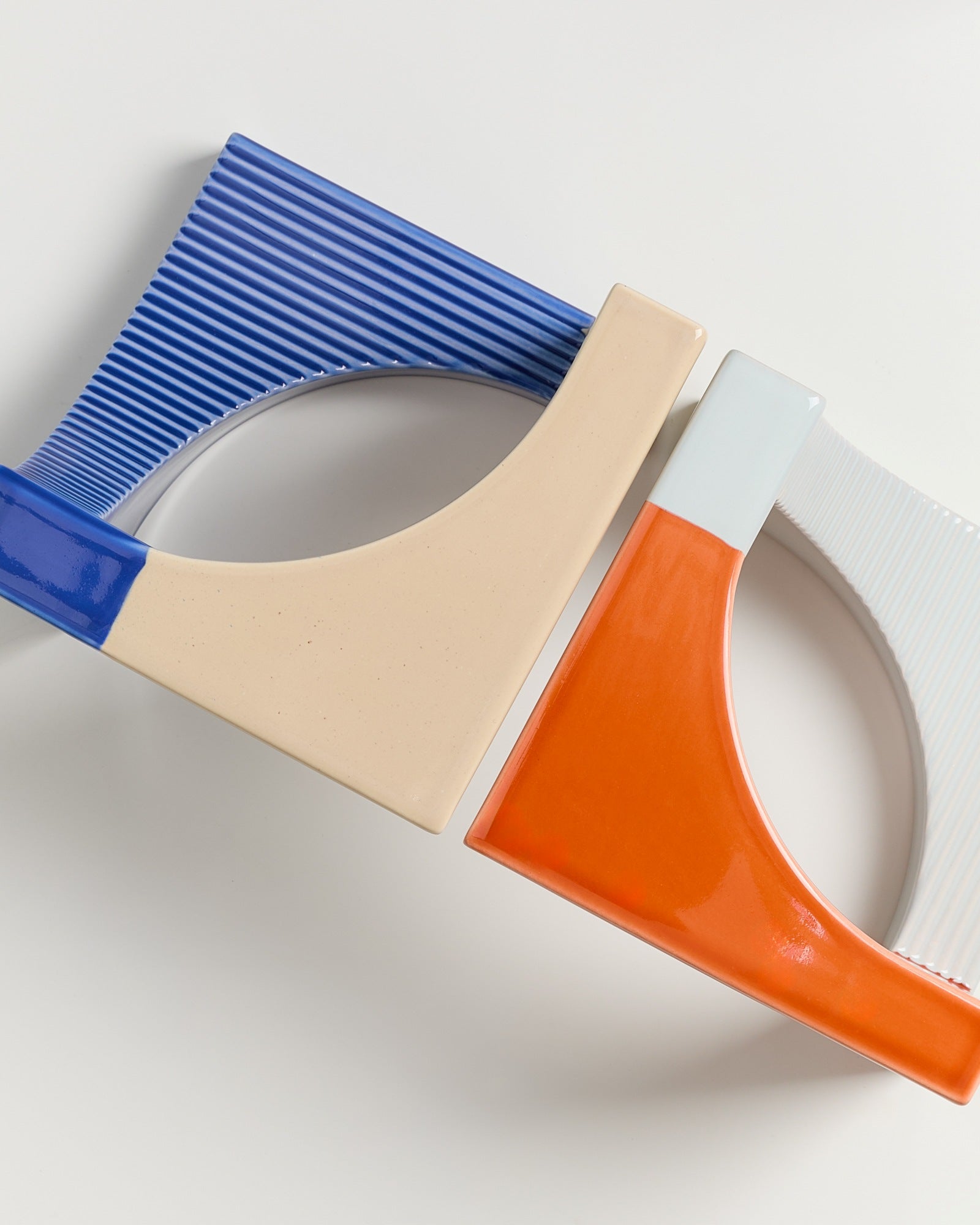 Zwei geometrische Keramikobjekte in Blau-Beige und Orange-Weiß, die ineinander verschlungen auf einem weißen Hintergrund liegen.