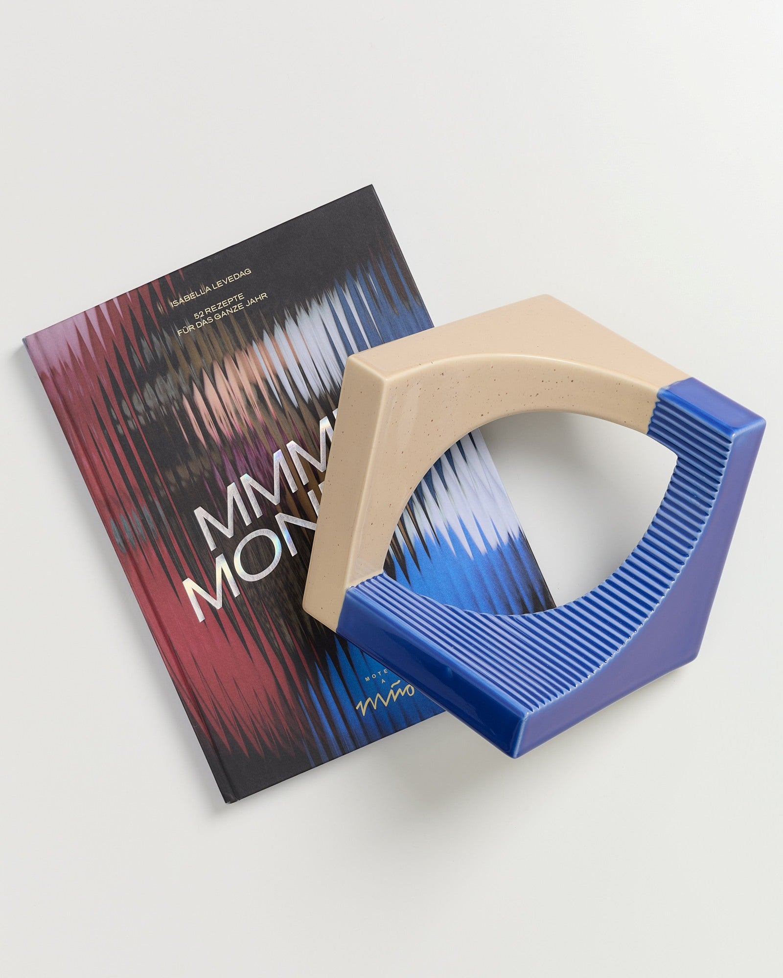 Ein Buch mit buntem, wellenförmigem Cover liegt unter einem geometrischen, beige-blauen Dekorstück.