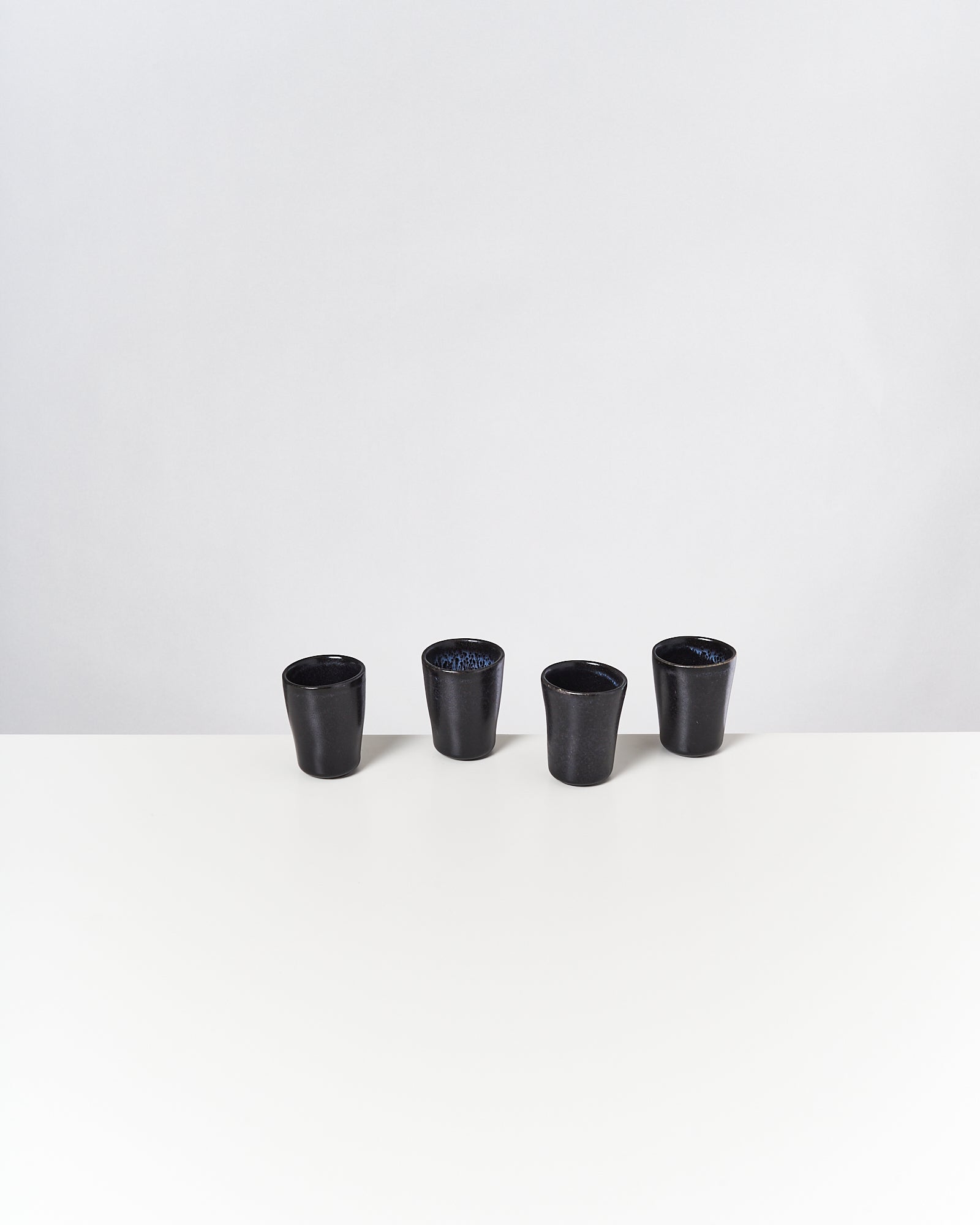 Vier schwarze Keramikbecher stehen auf einem weißen Tisch vor einem grauen Hintergrund.