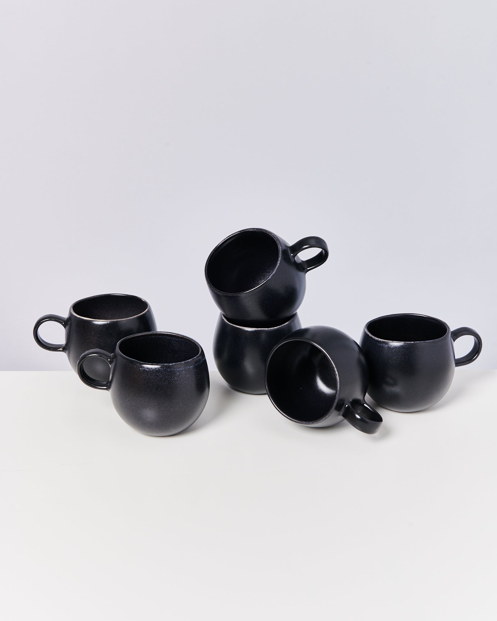 Sechs schwarze Tassen, von denen zwei gestapelt sind, auf einem weißen Tisch vor einem grauen Hintergrund.