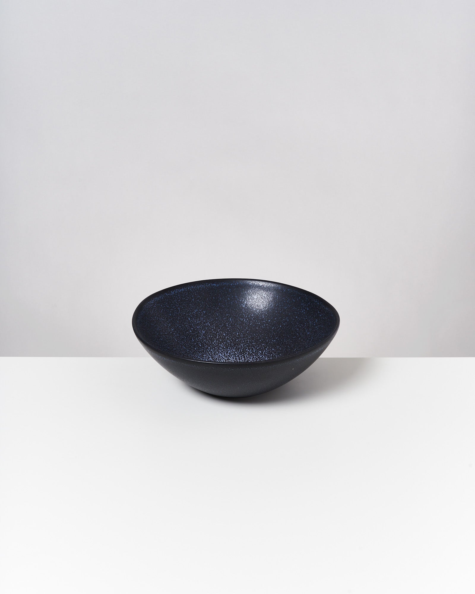 Dunkle, runde Schale mit strukturierter Oberfläche auf weißem Tisch vor grauem Hintergrund.