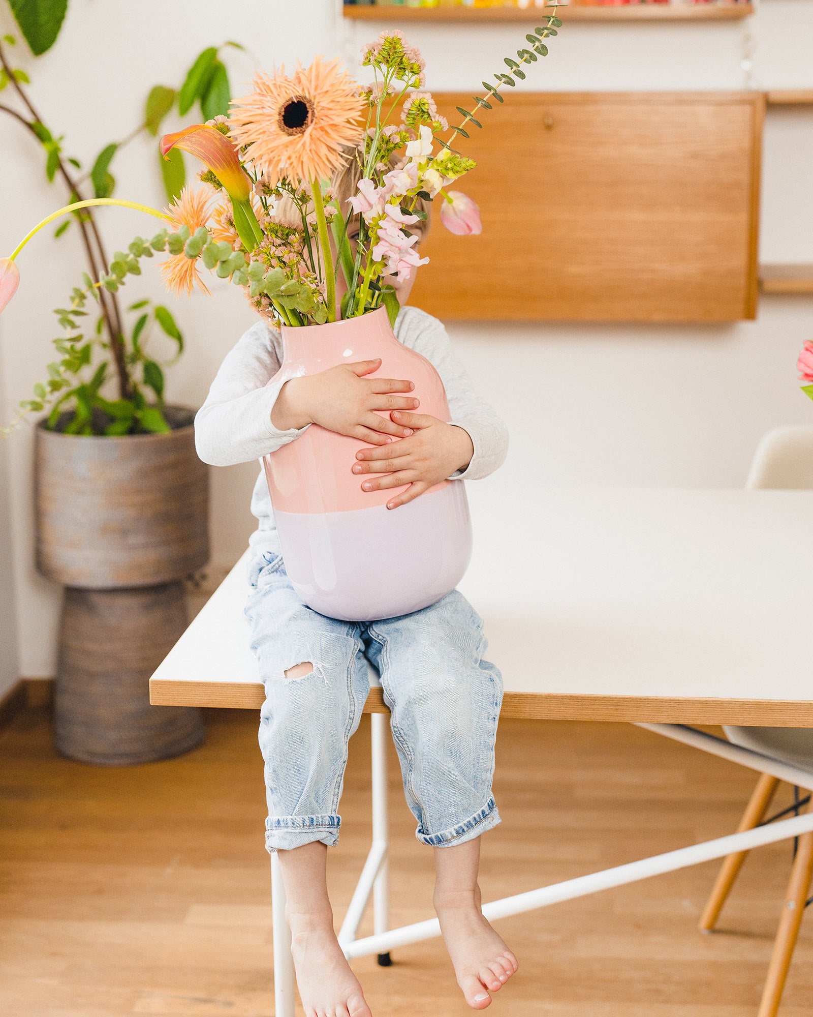 Kind sitzt auf Tisch und hält eine große Vase mit bunten Blumen, verdeckt sein Gesicht.