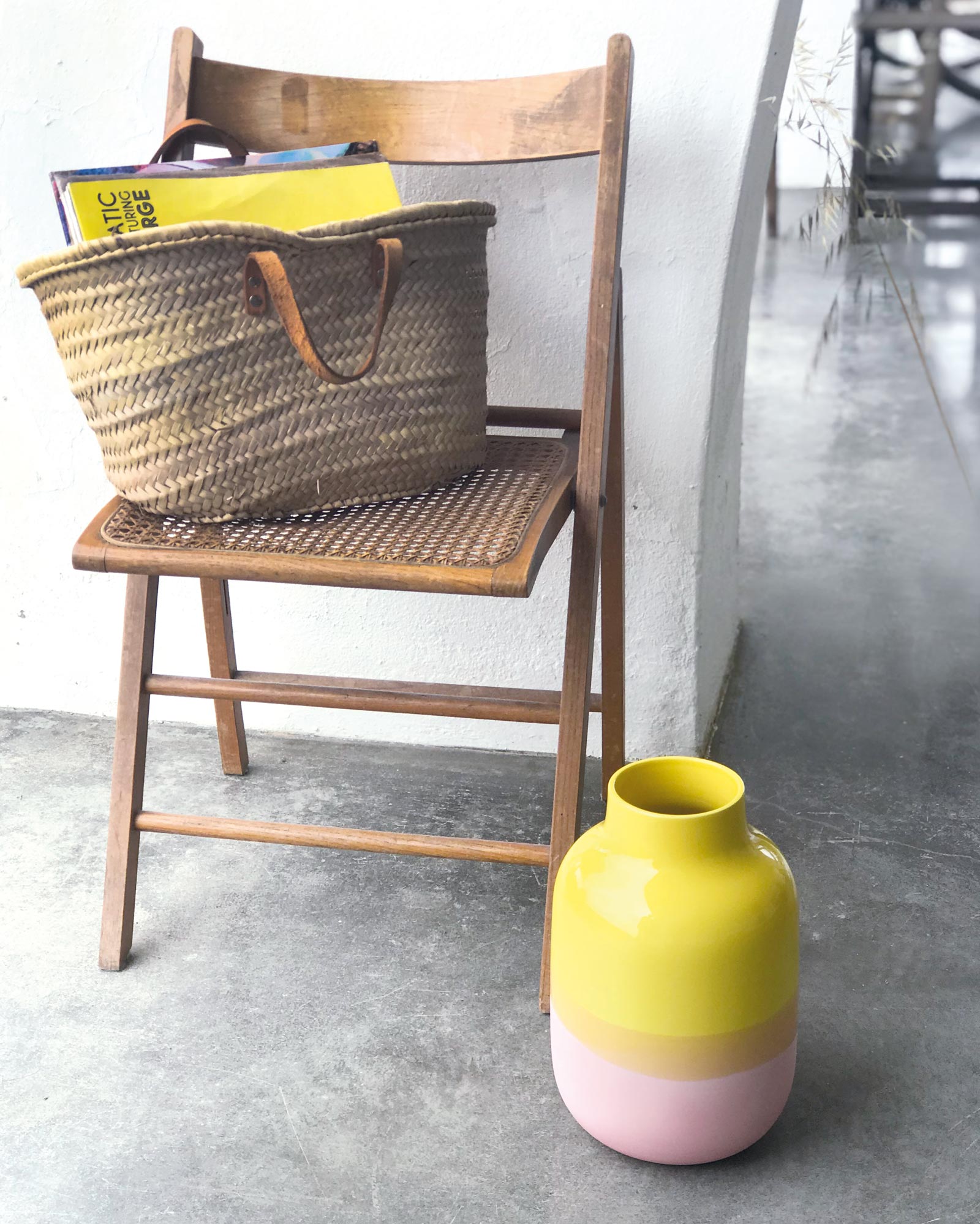 Ein Holzstuhl mit einem geflochtenen Korb darauf und daneben eine gelb-rosa Vase auf einem Betonboden.