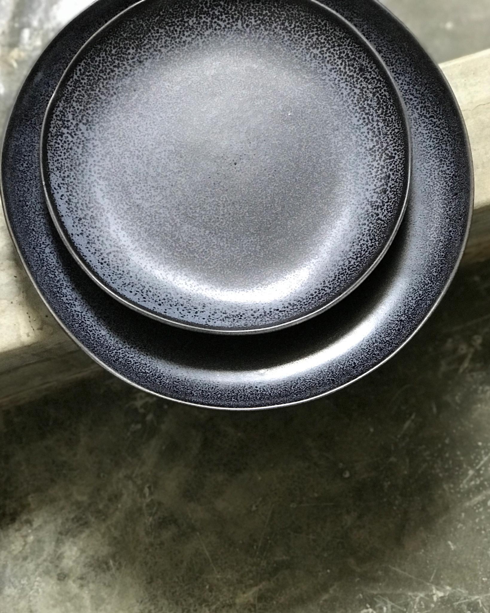 Zwei schwarze, körnige Teller liegen übereinander auf einem grauen Betonuntergrund.