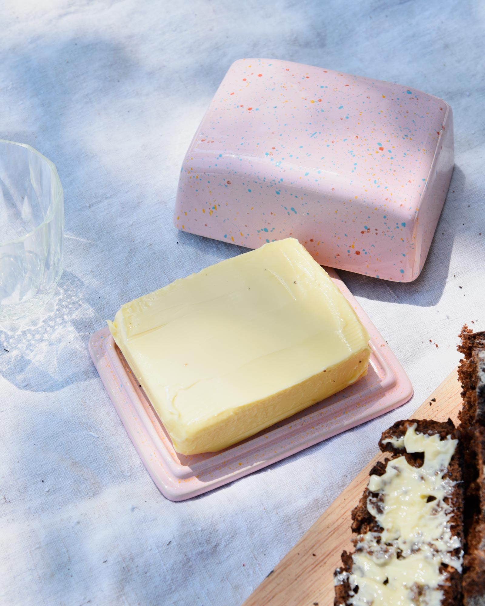 Ein Stück Butter auf einer rosa Butterdose mit Streuseln und bestrichenem Brot neben einem leeren Glas.