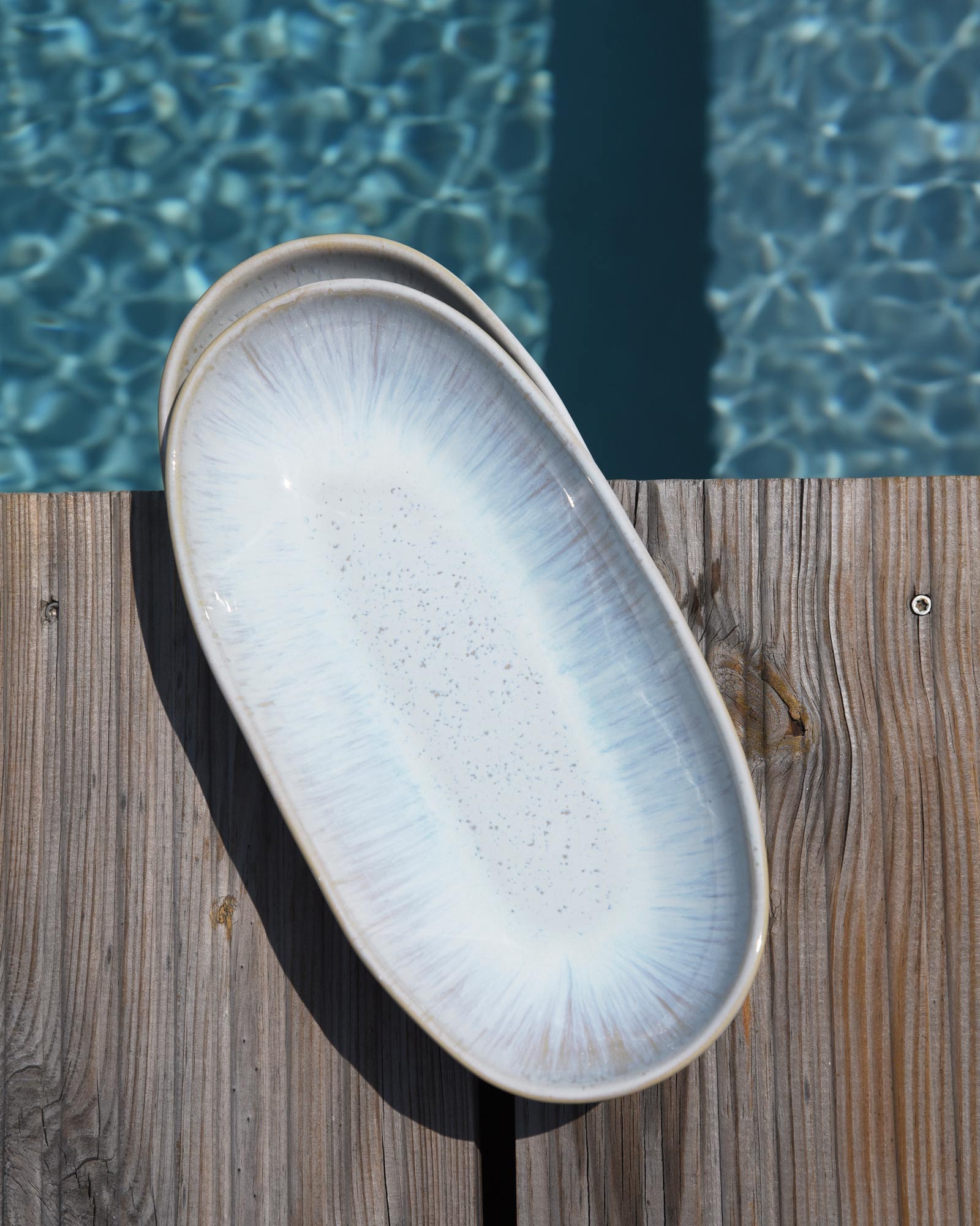 Ein ovaler, hellblauer Keramikteller liegt auf einem Holztisch neben einem Pool mit funkelndem Wasser.