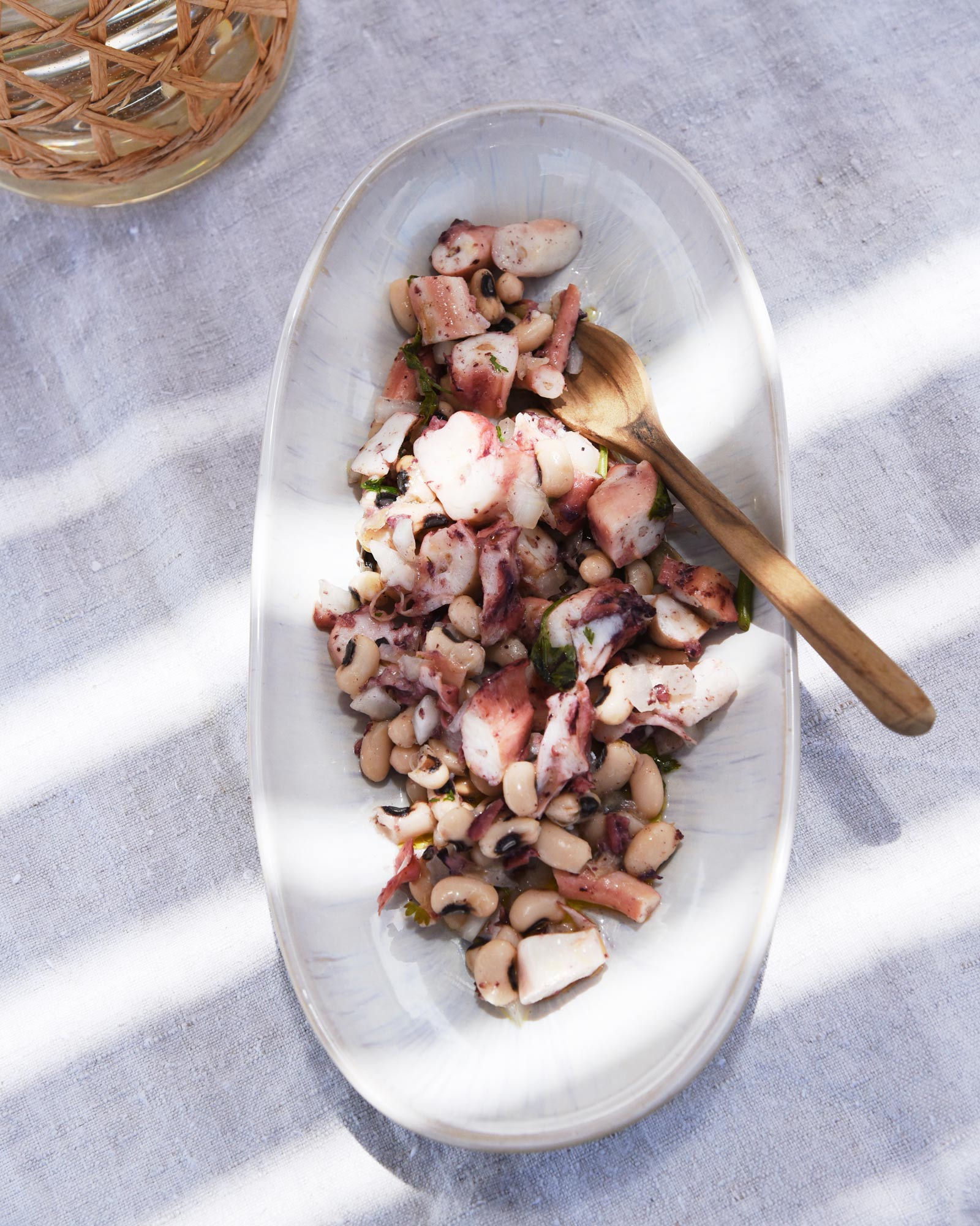 Oktapussalat mit Bohnen und Kräutern auf einem ovalen Teller mit Holzgabel, auf einem Leinentuch angerichtet.