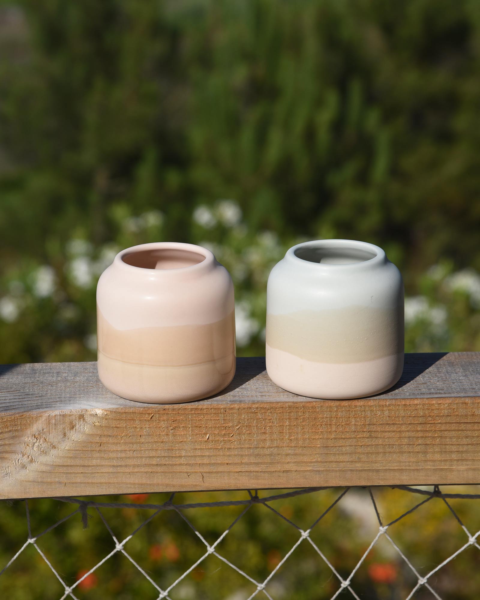 Zwei schlichte Vasen in Pastellfarben stehen auf einem Holzbalken vor grünem, verschwommenem Hintergrund.